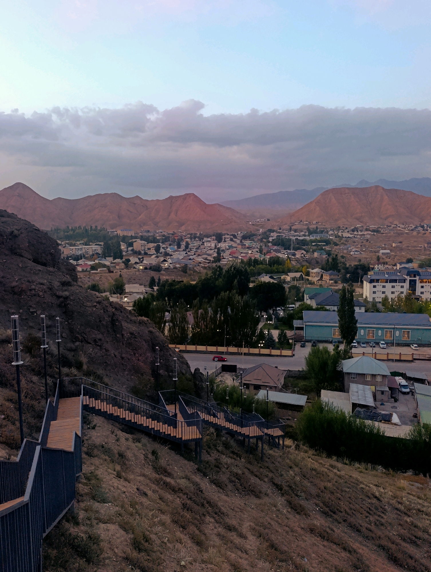 The image captures an elevated view of Naryn City, Kyrgyzstan, during what appears to be dusk or early morning. In the foreground, a steep, dry hillside is dotted with sparse vegetation. A modern, dark metal staircase with wooden steps winds down the slope, leading towards the city. The staircase is equipped with metal railings and intermittent modern light fixtures.
Beyond the hillside, the city of Naryn sprawls across a valley. Buildings of varying sizes and architectural styles, including residential homes and larger institutional structures, are visible. Some buildings have flat roofs, while others have pitched roofs. Trees are interspersed throughout the urban landscape, some appearing as clusters of darker foliage. A road runs through the city, with several cars parked or driving.
In the background, a range of arid, reddish-brown mountains rises, their peaks softened by the twilight sky. The sky itself is a gradient of pale blue at the top, transitioning to hues of pink and gray as it nears the horizon, with scattered clouds adding texture. The overall scene suggests a quiet, developing urban area nestled within a mountainous, semi-arid environment. No people are visible in the image.