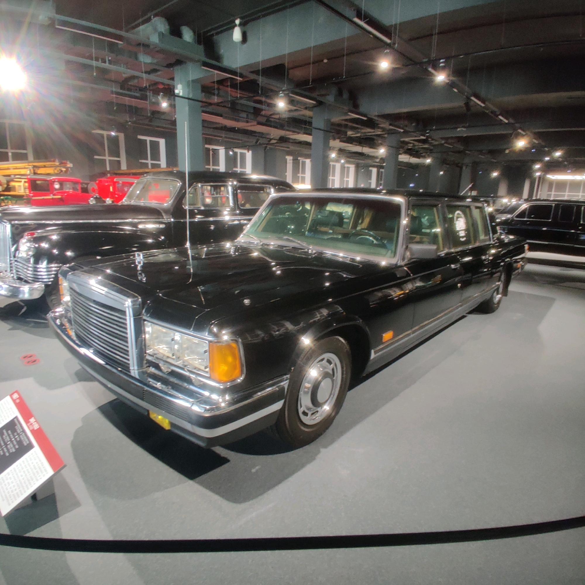 In the Museum of Military Technology "Battle Glory of the Urals" in Verkhnyaya Pyshma, Russia, a black ZiL-41047 limousine is displayed indoors. The limousine, manufactured in 1987, is positioned on a grey floor with a polished surface reflecting the overhead lighting and ceiling structure. To the left of the limousine, a red fire truck and part of another red vehicle are visible. A white informational sign with red accents is placed in the foreground at a slight angle to the limousine. The environment is an exhibition hall with dark ceilings, visible lighting fixtures, and structural elements. The limousine itself is the primary subject, showcasing its long body, chrome grille, headlights, and wheel. No people are present in the frame.