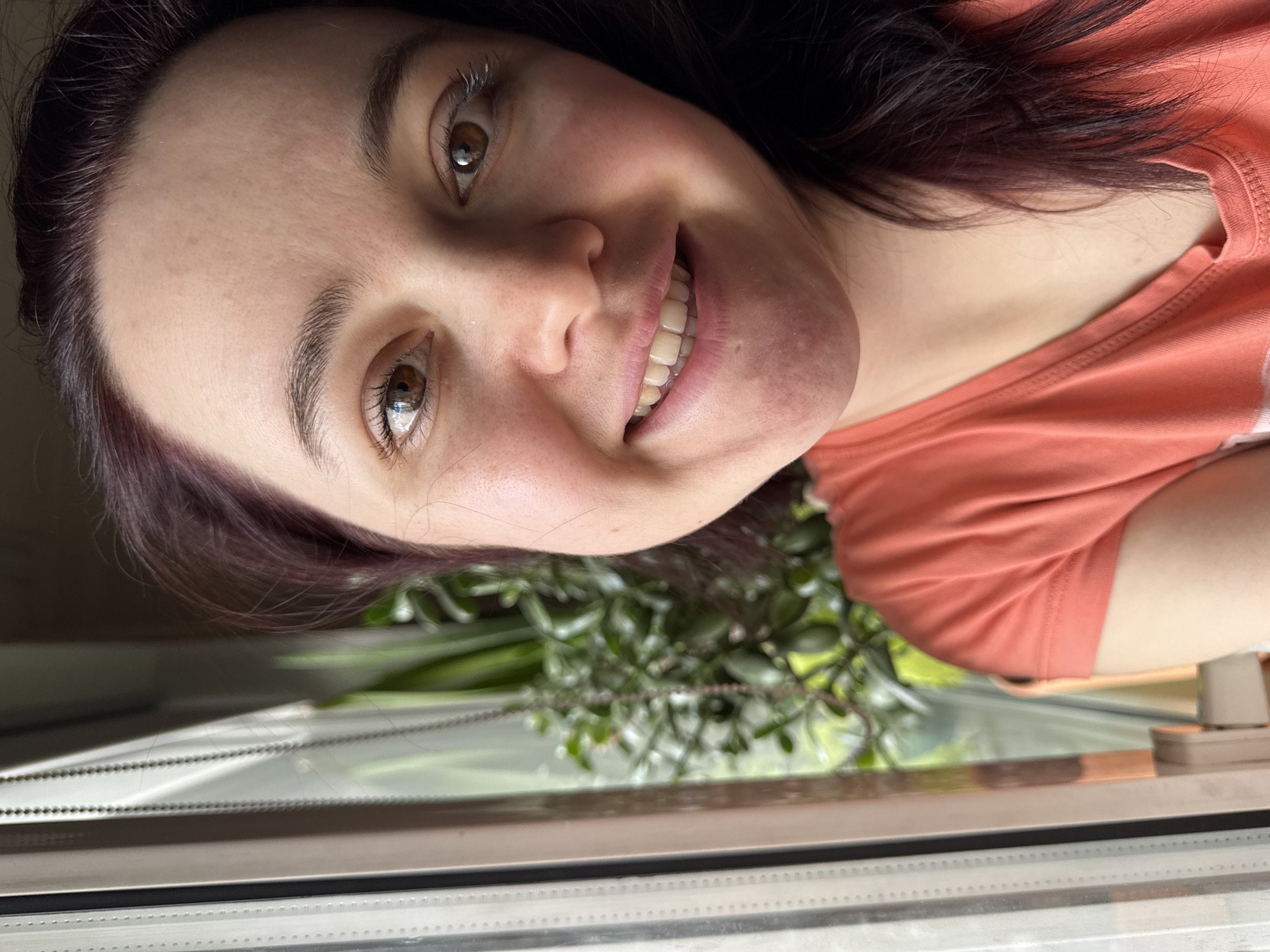 The image presents a close-up, rotated selfie of a smiling woman, taken indoors in Izhevsk, Russia. Her head is tilted significantly to her right, with her eyes looking upwards and slightly to the right, directly into the camera. She has dark hair, possibly with a subtle reddish tint, and warm brown eyes. Her face is illuminated by natural light, highlighting her clear skin, which shows natural texture around her nose and cheeks. She wears a coral or peach-colored t-shirt. A broad, friendly smile reveals her teeth, conveying a sense of joy and approachability. In the background, partially out of focus, is a window through which green foliage is visible, suggesting an outdoor garden or trees. The lower part of the window frame, including its sill, runs horizontally across the bottom of the image as it is currently oriented. The setting indicates daytime. No visible text is present.