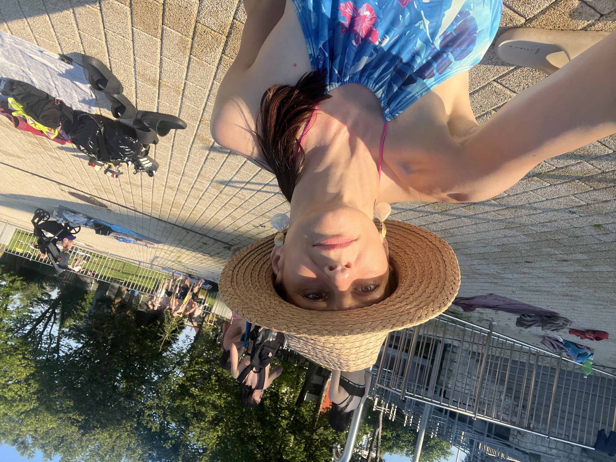 The image shows a woman wearing a straw hat and a blue swimsuit with a floral pattern. She is taking a selfie by a pool or park area. In the background, there are other people, trees, and a fence. There are also several items of clothing and shoes scattered on the ground. The sun is shining, casting shadows on the paved ground. The scene appears to be outdoors, possibly at a public swimming area.