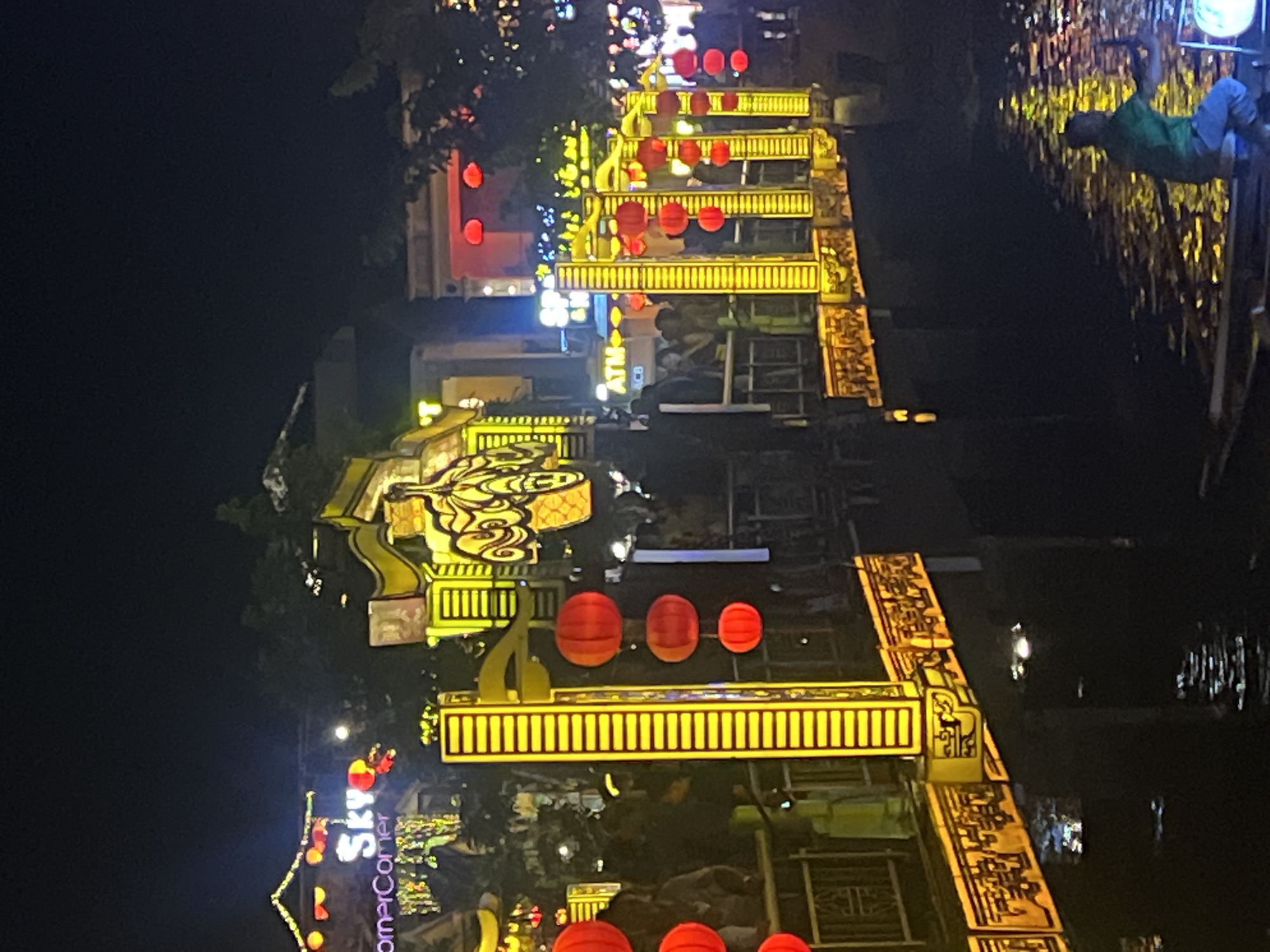 The image captures a nighttime view of a decorated bridge over a body of water in Hoi An, Vietnam. The bridge is illuminated by numerous bright yellow lights and adorned with red lanterns. The architectural style suggests traditional Vietnamese design, possibly within the Hoi An Ancient Town. Several people are visible on the bridge, some appearing to be tourists and others locals. The water reflects the vibrant lights, creating a shimmering effect. In the foreground, a person is seated in a boat, rowing on the water, with a round, glowing object in the boat. The scene conveys a festive and lively atmosphere characteristic of Hoi An's night markets and cultural events.