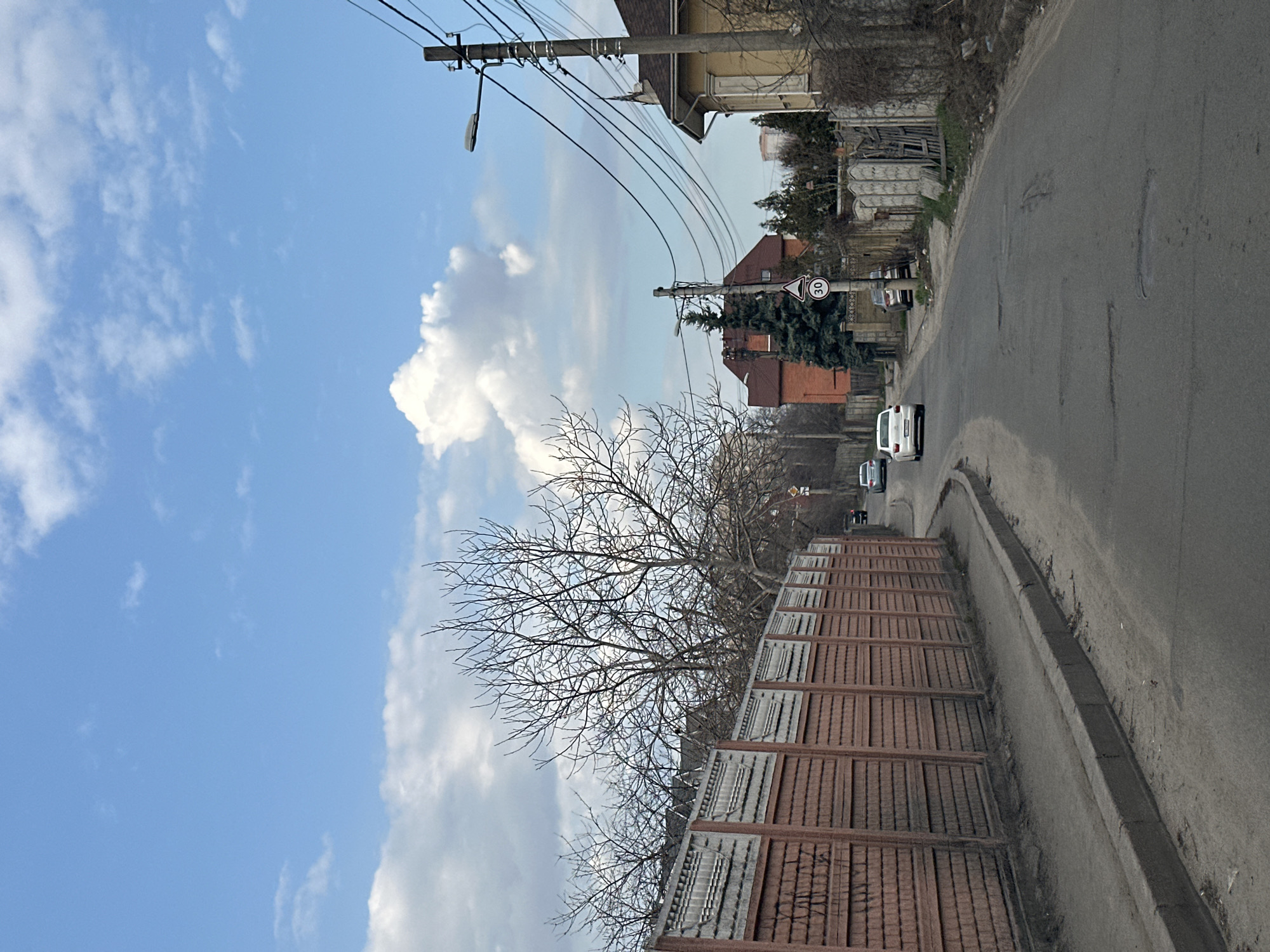 The image shows a street scene in Kyiv, Ukraine. The view is looking down a slight incline of a paved road with some potholes. On the right side of the road is a concrete curb and a low concrete retaining wall. On the left, a tall wooden fence runs along the sidewalk. Bare trees are visible above the fence. Several white cars are driving along the road in the distance. Utility poles with wires run along the left side of the road, and a street lamp is visible. Some buildings are visible further up the road. The sky is blue with scattered white clouds. A speed limit sign (30 km/h) is visible on the side of the road.
