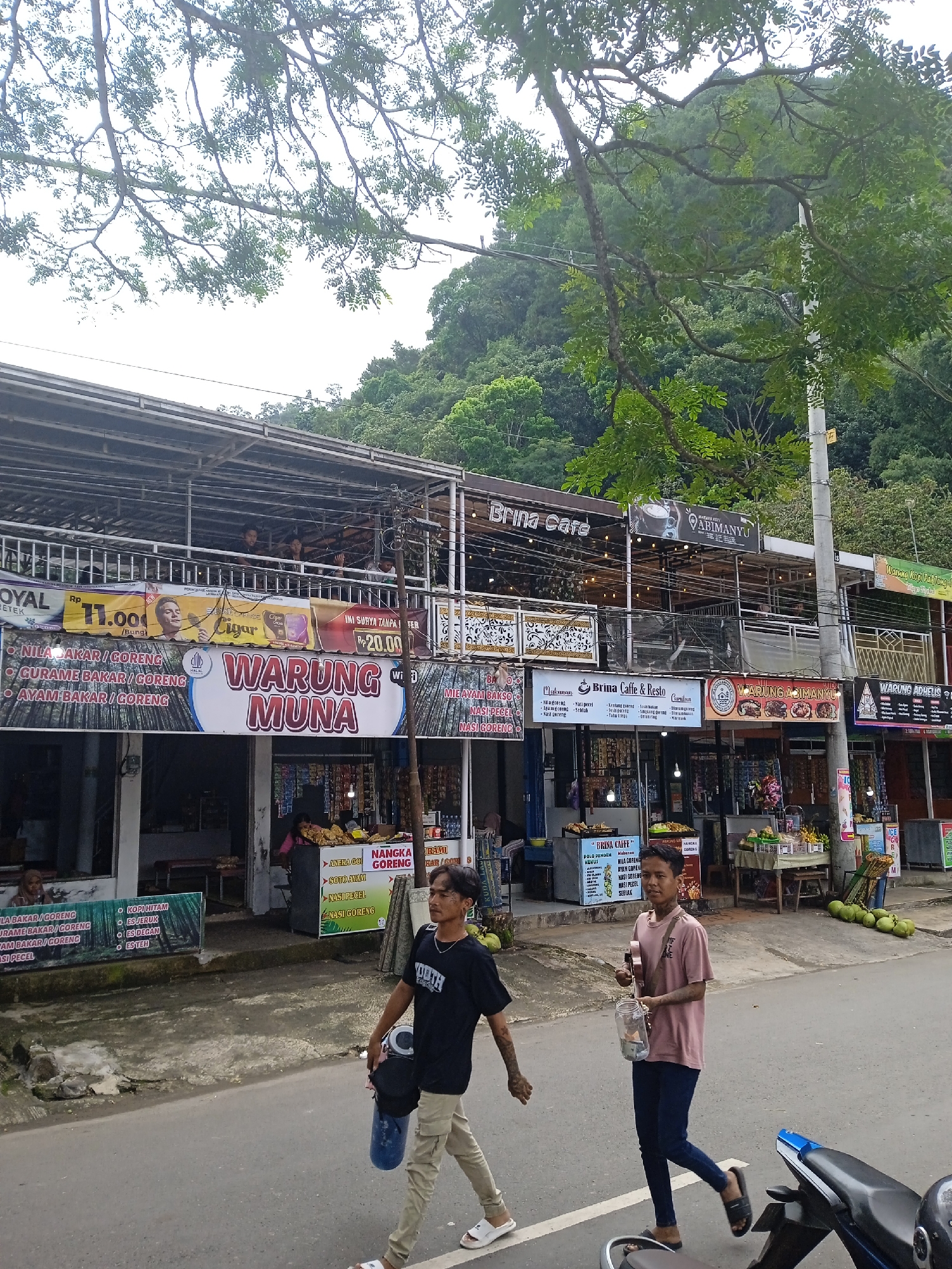 The scene is an outdoor street view in Nglingi, Indonesia. Several businesses line the street, mainly restaurants and cafes. They are multi-level with dining areas on the upper floors. Signage for businesses like "Warung Muna," "Brina Cafe," and "Warung Abimanyu" is visible. A couple of men are walking along the road, one carrying a blue container. A motorcycle is partially visible in the foreground. The area is backed by a forested hillside. The weather appears overcast.