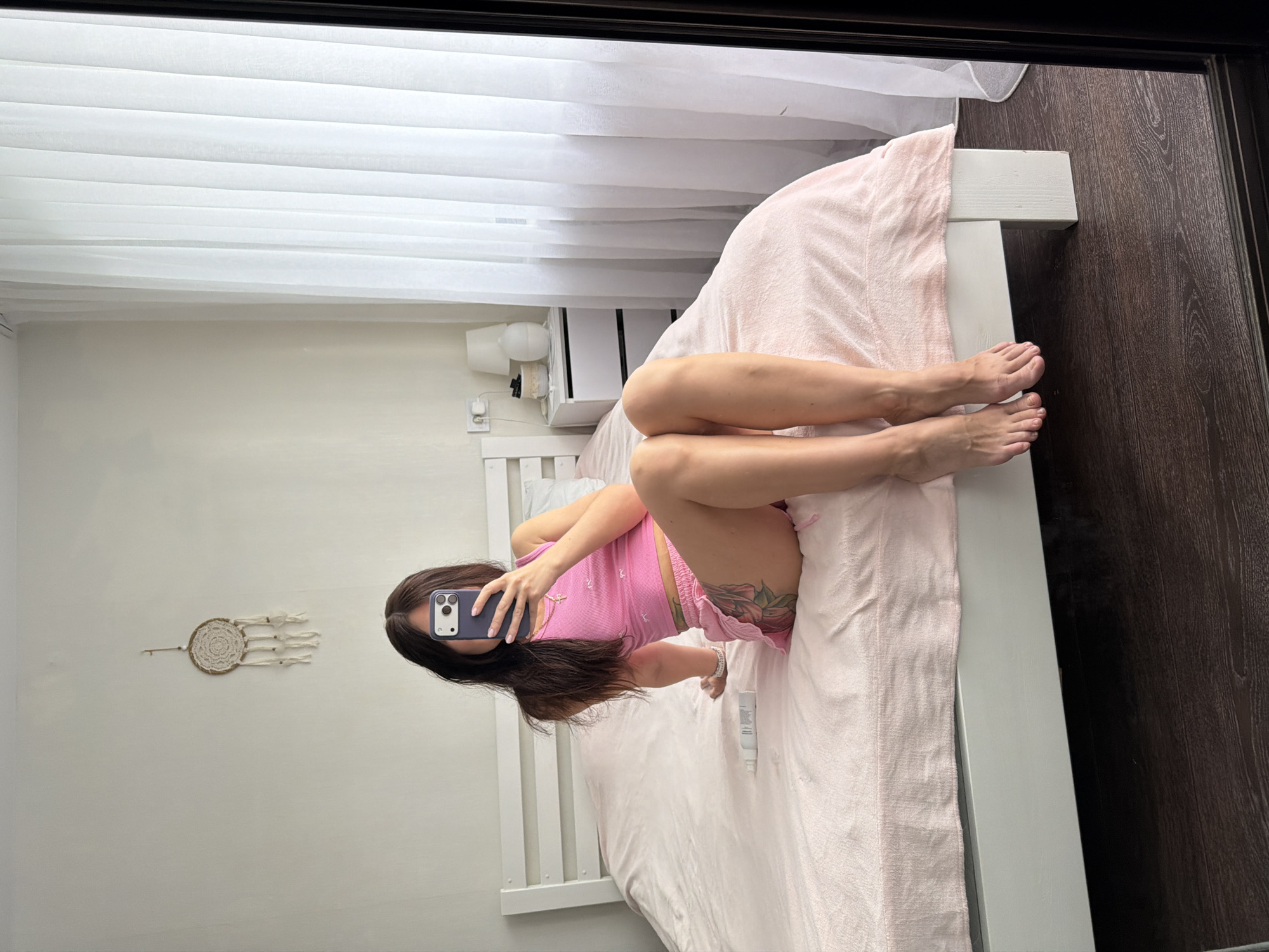A young woman is sitting on a bed in a bedroom. She is wearing a pink crop top and pink shorts. She is holding a phone in her right hand and taking a selfie. The bed is covered in a pink blanket. There is a nightstand next to the bed with a lamp and a diffuser on it. There are white curtains in the background. The room appears to be in an apartment in Sukhodol, Russia.