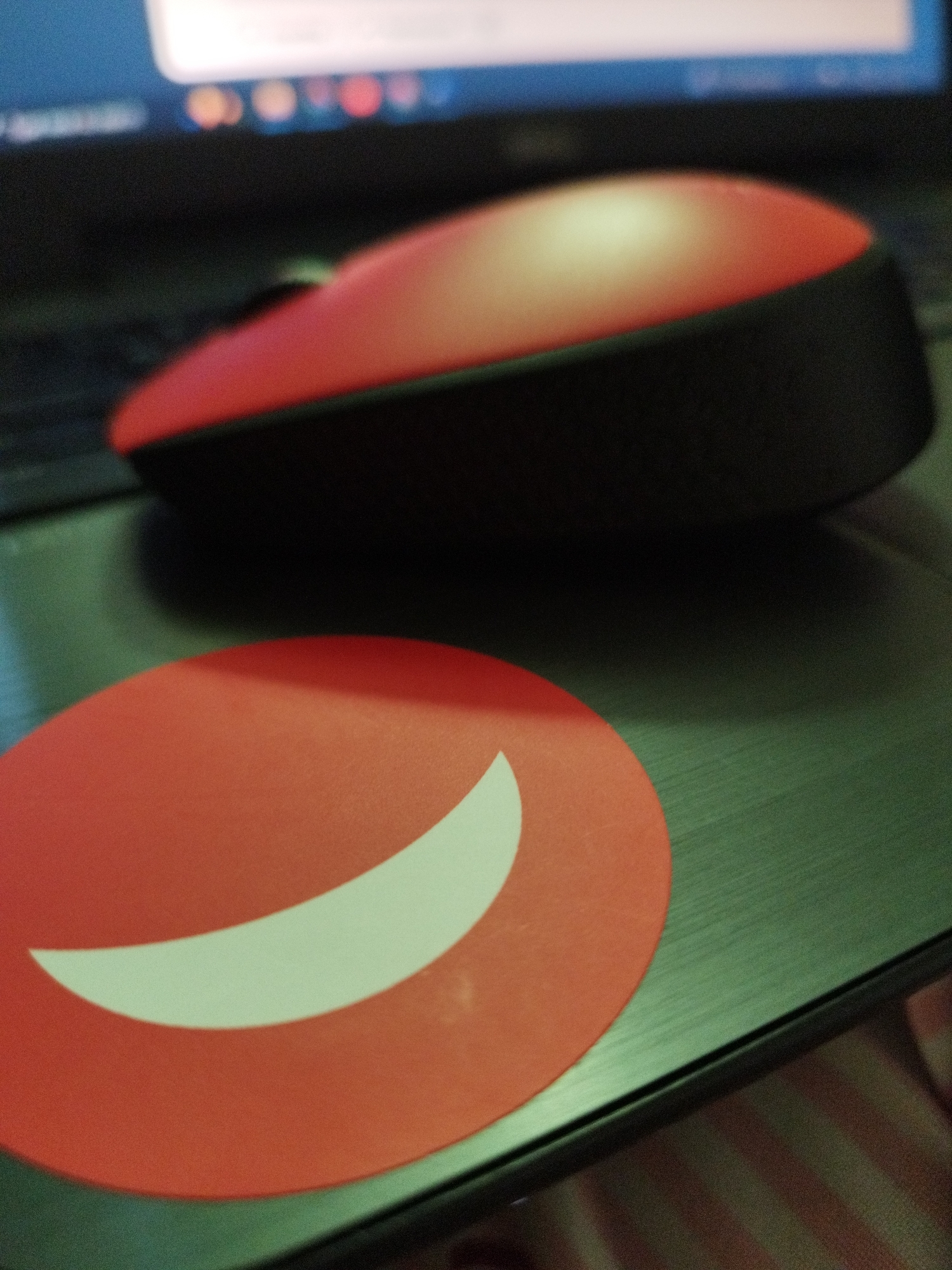 A red wireless mouse sits on a laptop next to a round, red sticker with a white crescent shape on it. The laptop screen is visible in the background, showing blurred images. The laptop surface is dark gray. Part of a striped pattern is visible in the lower right corner, possibly part of clothing or furniture. There is no information in the image to indicate that it is taken in Sofia, Bulgaria.