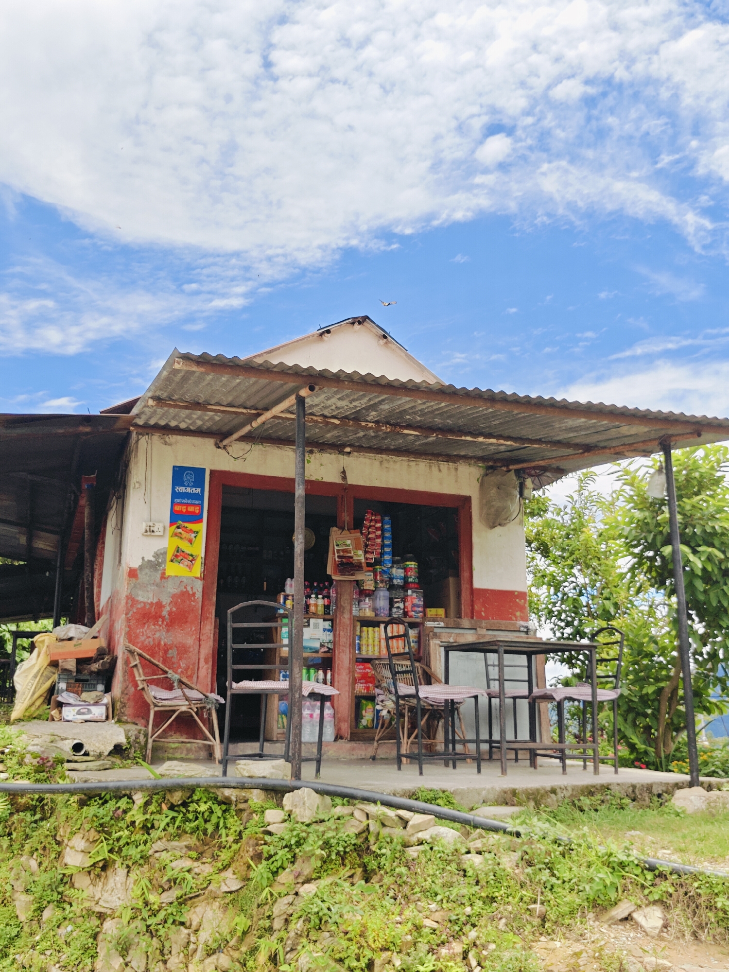This image depicts a small, open-air storefront in Pokhara, Nepal. The structure is built with a corrugated metal roof supported by metal poles, extending to cover an area in front of the shop. The walls are painted white and red, with some peeling and wear visible. Various items are displayed on shelves outside the shop, including bottles of drinks and packaged goods. In front of the shop, there is a small metal table with four chairs. A wooden chair with a cushion is placed to the left of the entrance. The storefront is situated on a slightly elevated platform, with a steep, rocky slope covered in greenery and vegetation below. The sky above is blue with scattered white clouds. The overall impression is of a humble, local business in a somewhat rustic setting.