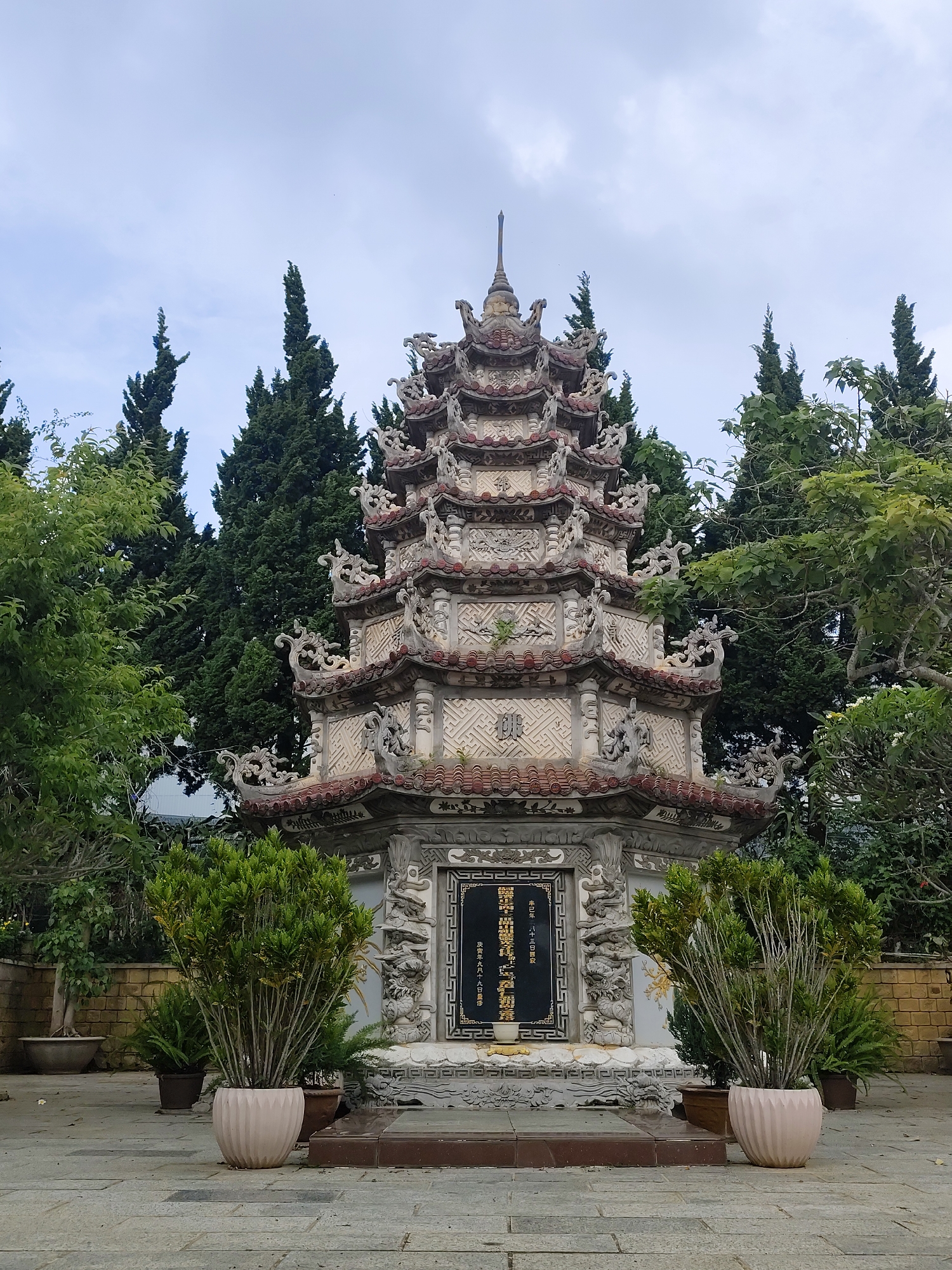 The image shows a multi-tiered pagoda-like structure, situated in what appears to be the grounds of a temple or garden, likely in Cam Ly - Đà Lạt, Vietnam. The structure has intricate detailing, with tiled roofs and ornate carvings. A stone plaque with inscriptions in vertical columns is embedded in the base. Tall coniferous trees are visible in the background, along with other greenery and potted plants flanking the structure. The ground is paved with stone tiles. The sky is overcast. There are no people visible in the image.