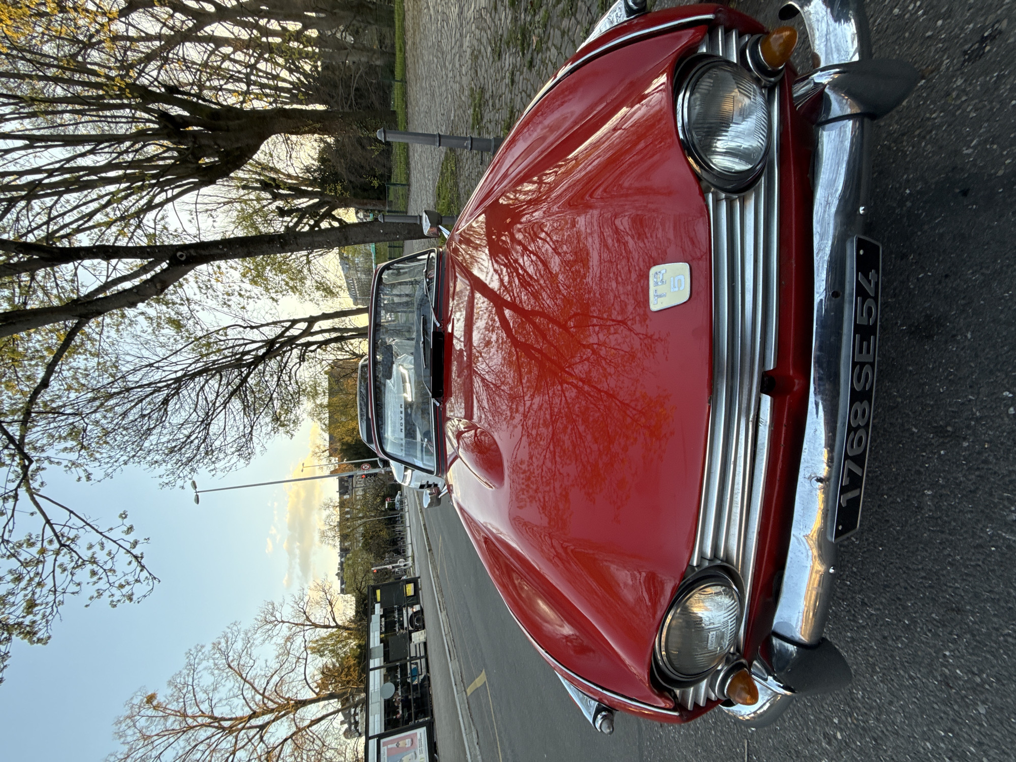 A red classic Triumph TR5 convertible is parked on an asphalt street in Strasbourg, France, facing the viewer. The car features a chrome front bumper, grille, and circular headlights, with an amber turn signal positioned near the left headlight. A black license plate displaying "1768 SE 54" is mounted on the front bumper. The "TR5" emblem is visible on the red hood, which reflects bare tree branches and the sky. The convertible top is down.
To the right of the vehicle, a cobblestone pathway runs alongside a grassy area with numerous deciduous trees, some appearing bare, others showing early signs of foliage. Gray bollards separate this area from the street.
In the background, on the left side of the street, a bus stop with advertising posters is visible. A white articulated bus is present near the stop, with several indistinct figures visible inside. Further down the street, a white Range Rover is partially visible behind the Triumph. Buildings with multiple stories line the distant street, and streetlights are intermittently placed. The sky above is light blue with scattered clouds, indicating a late afternoon or early evening setting. No individuals are clearly discernible interacting with the scene or the primary vehicle.
