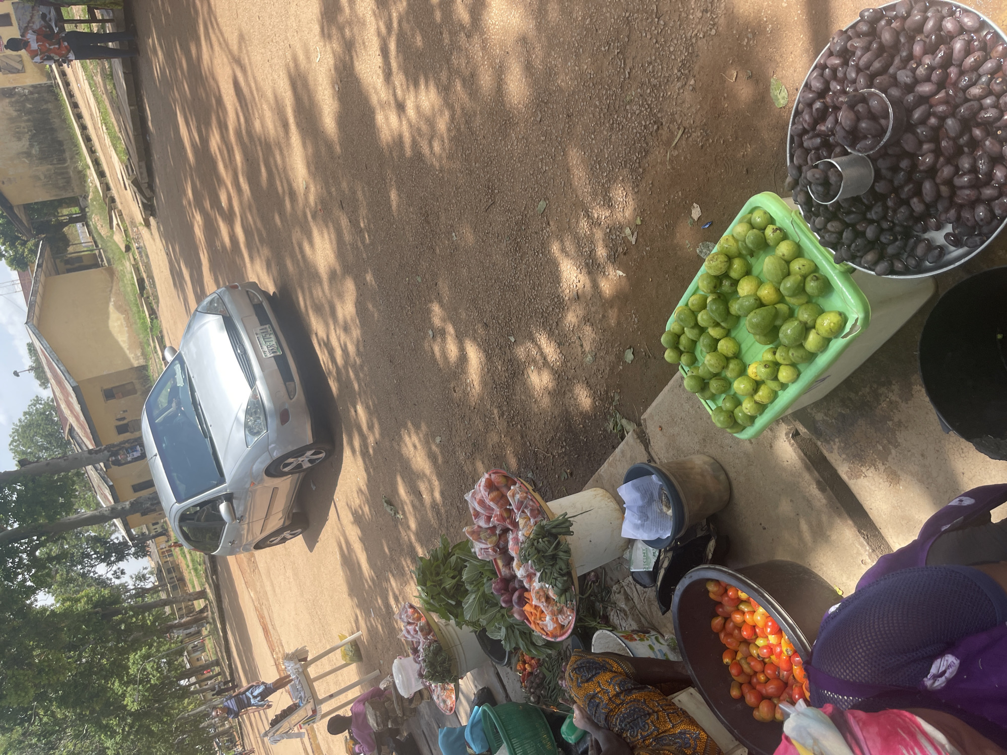 This image captures an outdoor market scene in Rafin Par, Nigeria. A silver sedan is parked on a dirt road, which is partially shaded by trees. In the foreground, several stalls display fresh produce. One stall features a large metal basin filled with dark, oval-shaped fruits, likely olives, with a smaller metal cup also containing some of these fruits placed inside. Next to it, a bright green plastic container is overflowing with small, round, light green fruits, possibly unripe guavas. Further down, other stalls showcase baskets of vegetables and fruits, including tomatoes, onions, and leafy greens.
People are present, though their faces are not clearly visible. Some individuals are tending to the stalls, while others appear to be customers or passersby. One person is wearing a purple headscarf and a dark blue patterned garment. Another person is wearing a colorful patterned dress. The overall atmosphere seems to be that of a typical bustling market day. The sunlight filtering through the trees suggests it is daytime, likely mid-morning or afternoon. The presence of vehicles and market stalls indicates a populated area with commercial activity. The ground is covered in loose dirt and some scattered leaves. There is no visible text in the image.