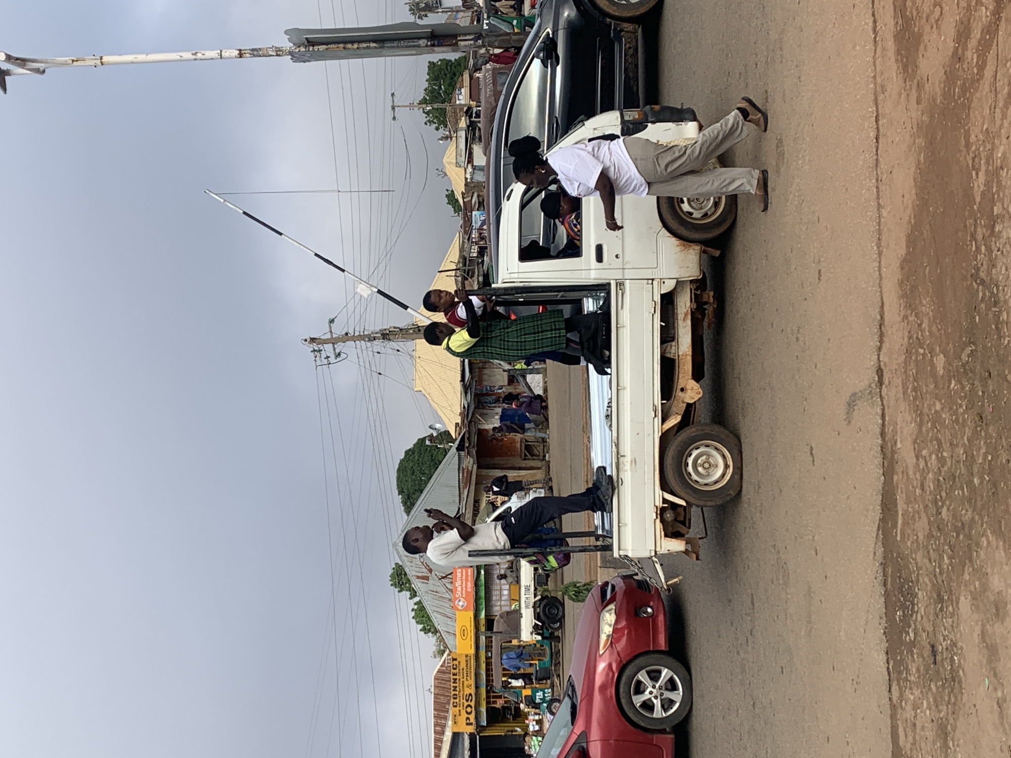 In Gura Top, Nigeria, a white flatbed truck is stopped on a paved road, possibly at a traffic crossing. A man is standing in the truck bed, facing left, holding a smartphone. Three young women are also in the truck bed; one in a green and yellow uniform is standing, facing the camera. To the right of the truck, a woman in a white shirt and light-colored pants is walking away from the truck, facing right. A black SUV is behind the truck, and a red car is to the left of the truck. Buildings line the street, with signs for "CONNECT POS & PHONES" and "StarTimes." A white pole with a striped barrier extends from the right side of the frame. The sky is overcast.
