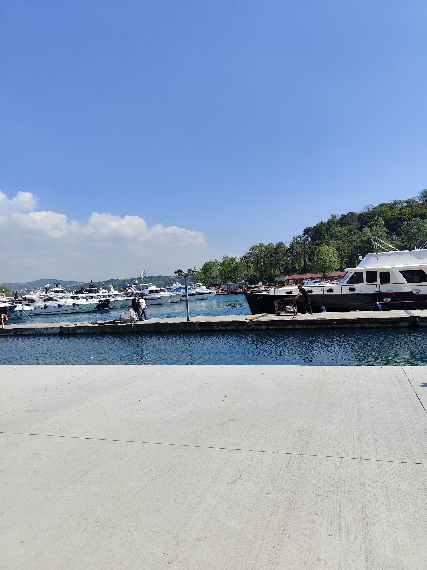 This image depicts a marina in Sarıyer, Turkey. The scene is captured on a bright, sunny day with a clear blue sky dotted with a few cumulus clouds. In the midground, there are numerous boats docked at a concrete pier. The water is a deep blue, reflecting the sky. Two people are visible walking on the pier among the boats. A man is seen tending to a boat on the right. A wooded hill rises in the background. The foreground is dominated by a wide, light gray concrete surface, likely part of the marina's walkway.