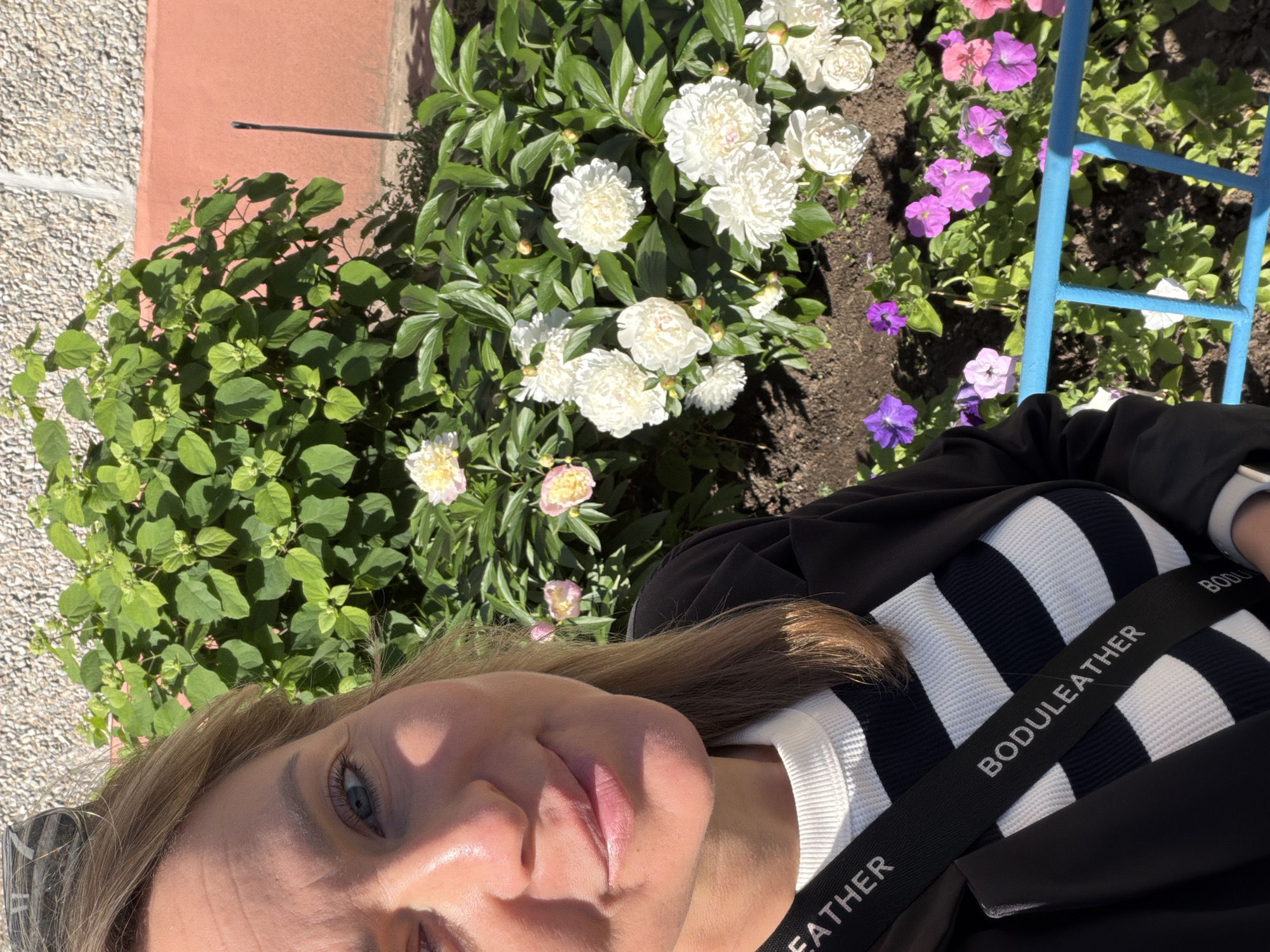 A woman is taking a selfie in front of a flower bed in Chaikovsky, Russia. The woman is wearing a black jacket over a white and black striped sweater, and a black strap with white text that reads "BODULEATHER" is draped across her chest. She has blonde hair and blue eyes, and she is smiling. The flower bed contains numerous white peonies and some smaller pink and purple flowers. Behind the flowers, there is a green bush, a textured wall, and a pink wall. The scene appears to be outdoors on a sunny day.