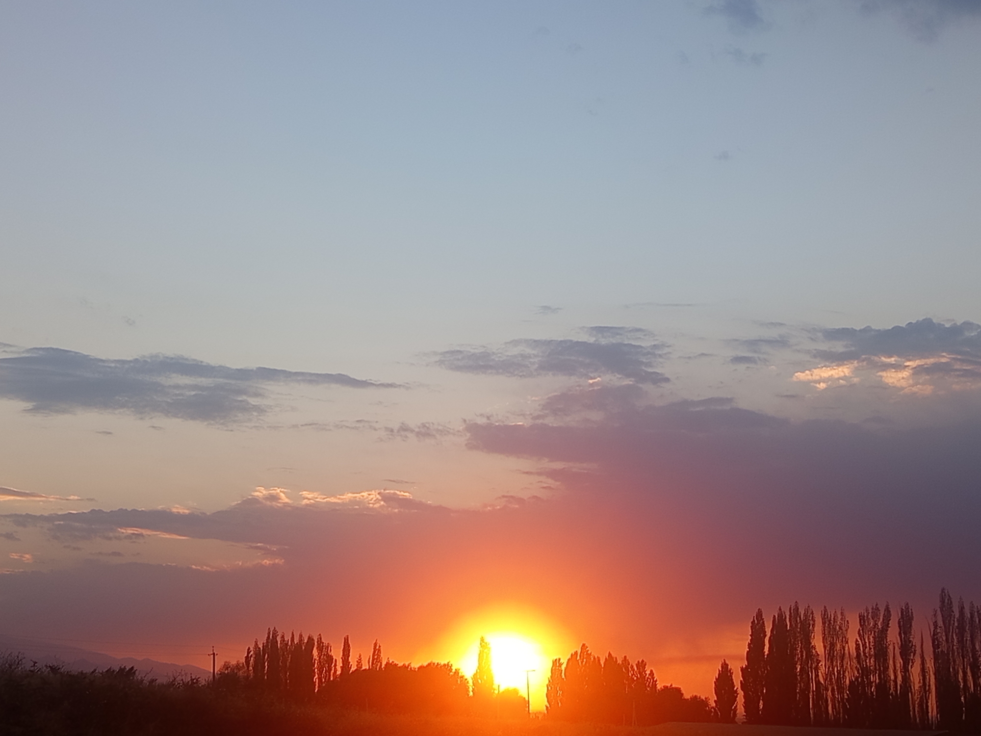 This is a wide-angle, eye-level, medium shot of a sunset over a rural landscape. The sky is a gradient of colors, from pale blue at the top to fiery orange and pink near the horizon. Clouds are scattered across the sky, some tinged with the colors of the sunset. Below the sky, a silhouette of trees and hills forms the horizon line. In the distance, the setting sun is visible as a bright, glowing orb, illuminating the sky with its light. The overall impression is one of natural beauty and tranquility. It is difficult to pinpoint the exact location in Jardy-Su, Kyrgyzstan, but the scene depicts a characteristic rural landscape with trees and hills. There are no people or specific objects that would indicate a particular landmark or activity within Jardy-Su.