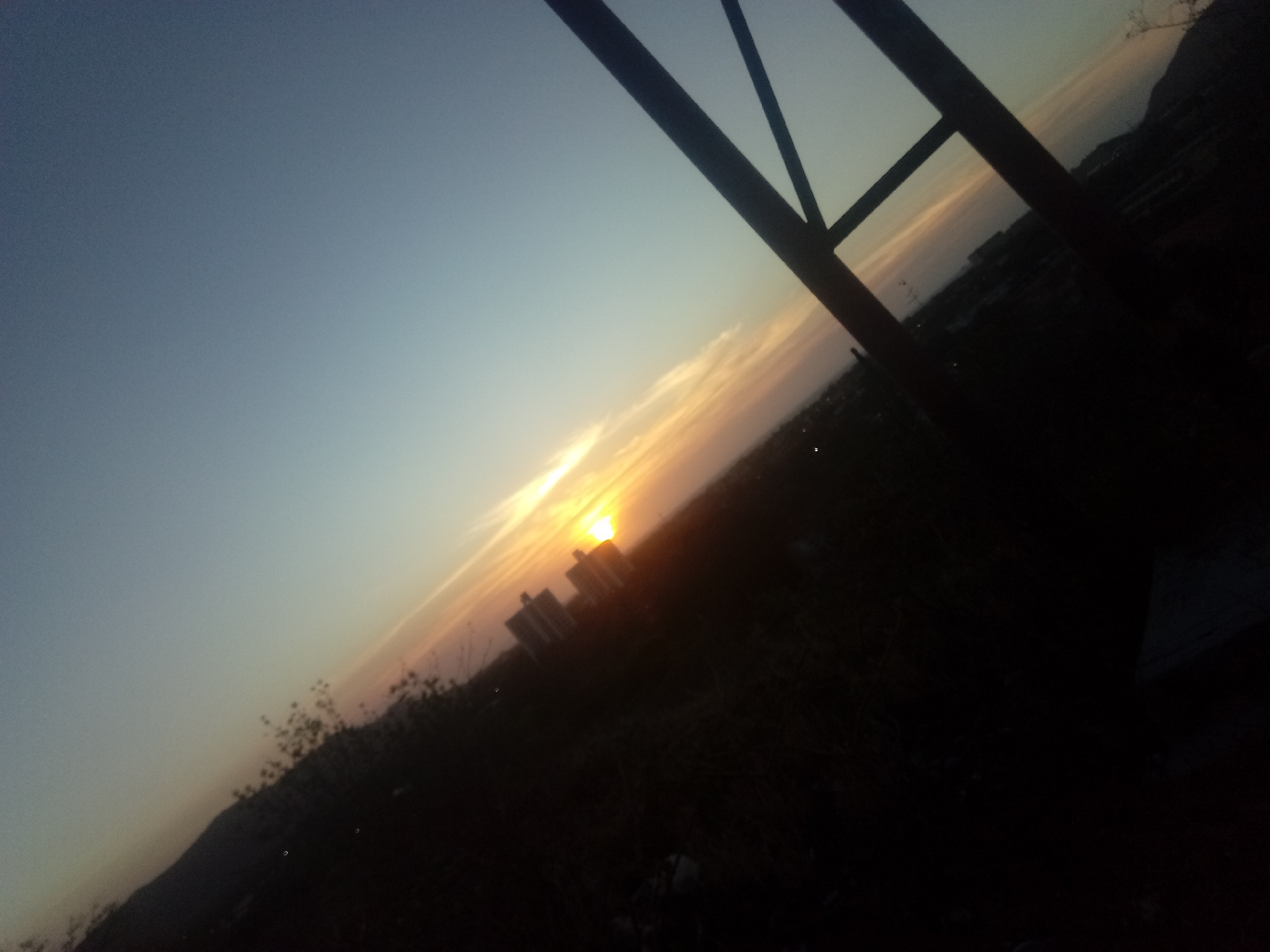 The image shows a cityscape at sunset, likely taken from an elevated position. The sun is setting behind buildings, creating a bright golden light streaking across the sky. The foreground is dark, with silhouetted vegetation and a metal structure partially obscuring the view. Given the request includes the city of Altagracia, Venezuela, it's plausible that the buildings in the image are located in that city. However, without further information, it is impossible to confirm the location precisely. There are no discernible people or actions visible in the image.
