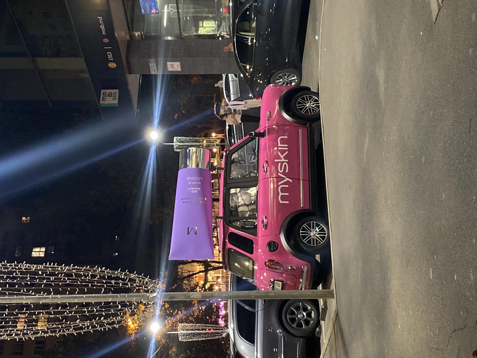 A pink, compact car with a large purple tube of skincare product on its roof is parked on a street at night. The car has "myskin" written on its side. In the background, there is a building with illuminated windows, street lights, and other parked cars. A sign on the building reads "ma ib 365" and "maib.md". The scene appears to be in Chișinău, Moldova, as indicated by the "maib.md" domain and the likely presence of a "maib" bank branch. The overall atmosphere is urban and nocturnal.