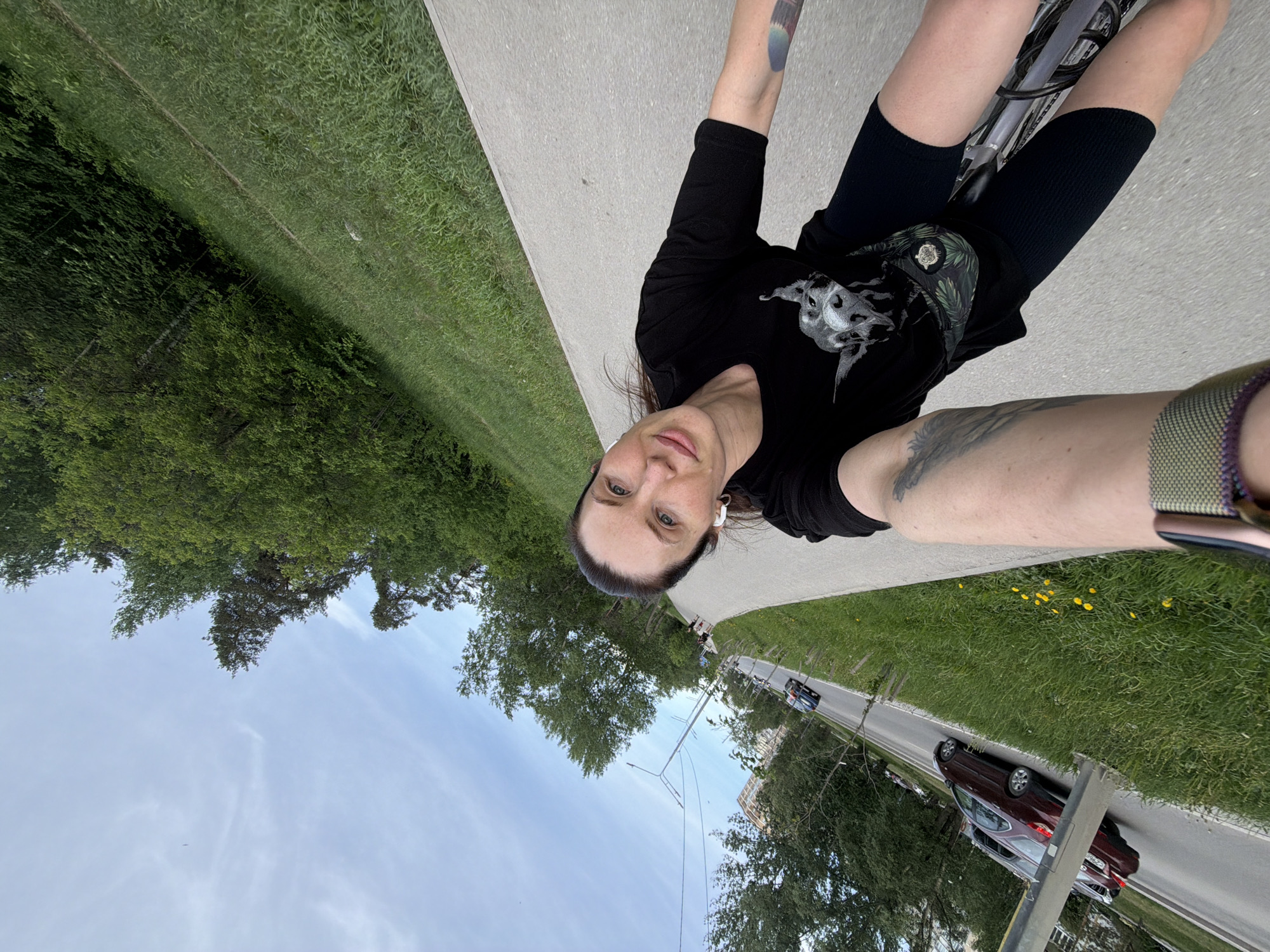 A woman is taking a selfie while riding a bicycle on a paved path. The path is bordered by grass and trees on one side and a road with cars on the other. The sky is visible in the background. The woman is wearing a black t-shirt and knee warmers. She has visible tattoos on her arms. The location is Perm, Russia.