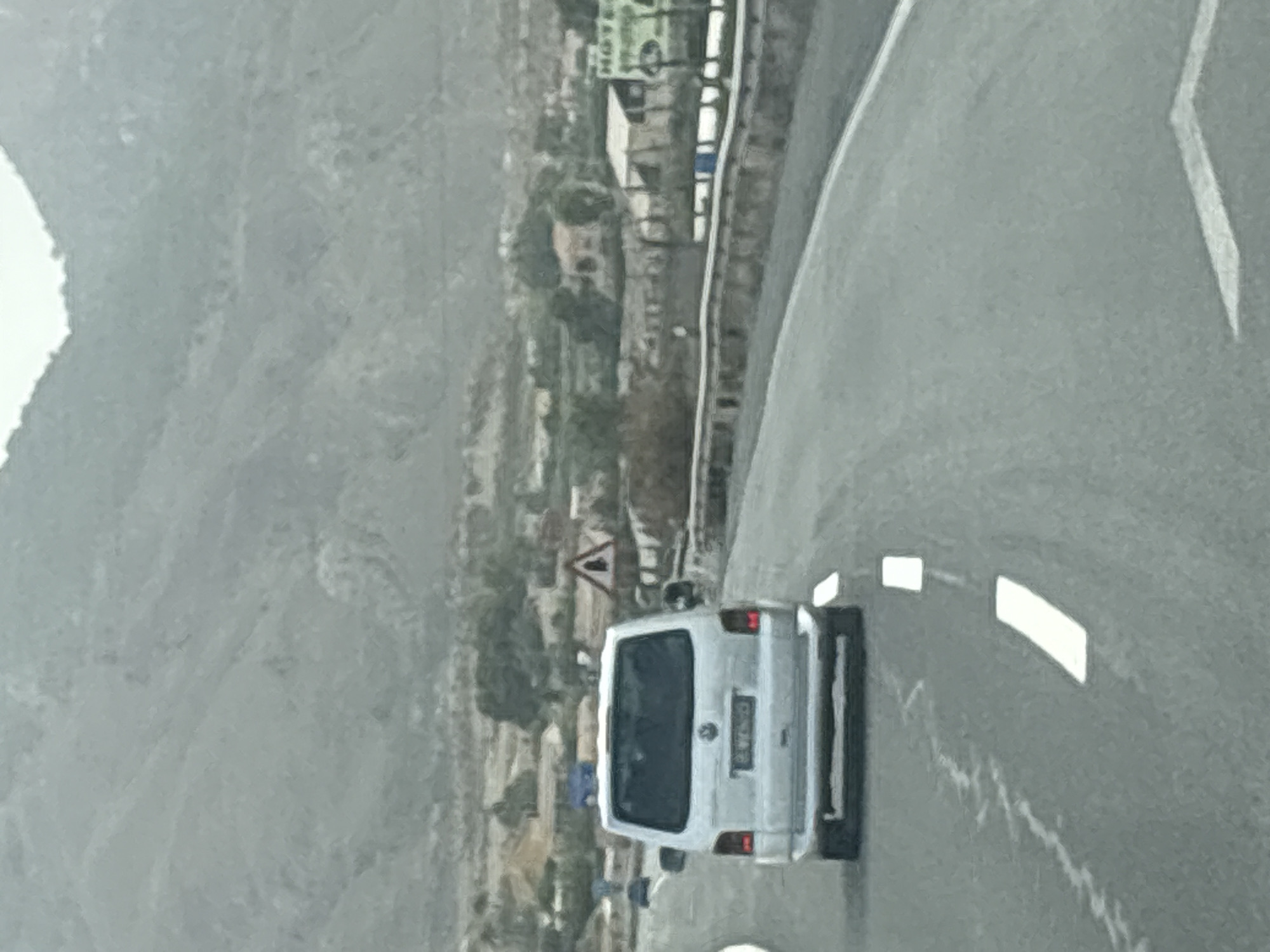 This media file shows a view from a vehicle driving on a road in Lorca, Spain. The main subject is a white van, seen from the rear. The van has a license plate with some visible text, but it is not clear enough to read. The van is traveling on a paved road with white lane markings. In the background, there are hills or mountains with a somewhat sparse, arid landscape. Some buildings and vegetation are visible on the slopes of the hills, suggesting a semi-rural or suburban setting. A triangular road sign is visible on the left side of the road. The sky appears overcast, and the overall lighting suggests it might be daytime, possibly in the morning or late afternoon, with diffused light. The image is taken from a low angle, looking forward and slightly to the left, as if from the passenger seat of a following vehicle. The road curves to the right.