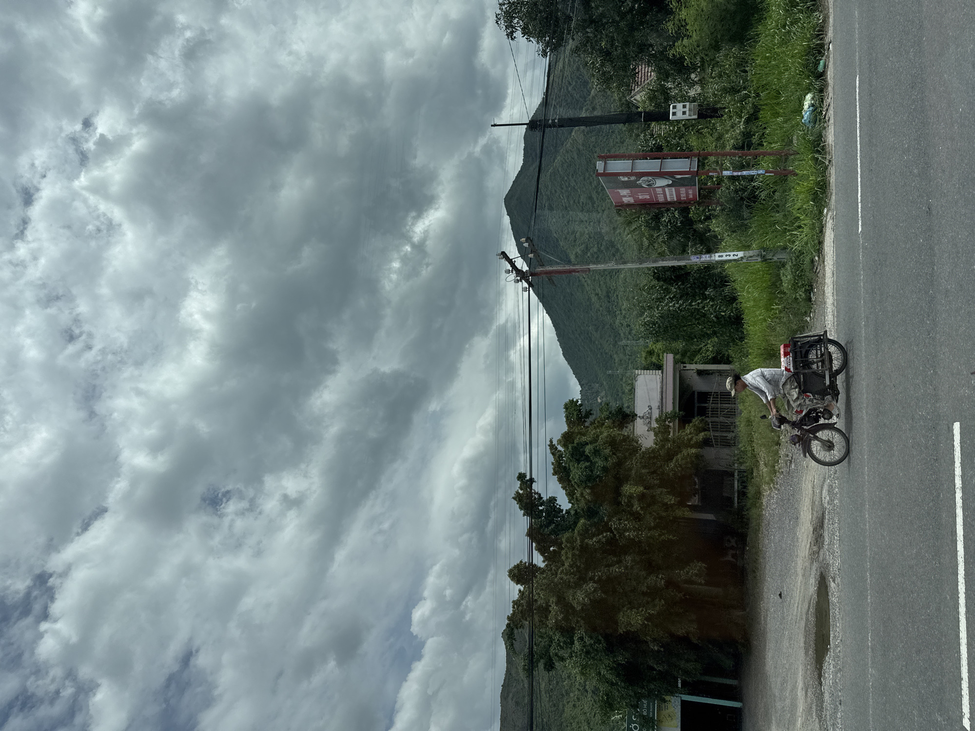 A person is riding a motorbike with a cargo rack attached to the rear. The motorbike is on the side of a paved road. The road has a white line marking the lane. The weather appears cloudy. The background features a lush green mountain covered in vegetation. There are power lines running across the scene. A red and white sign is visible near the road, with some text in a foreign language. The overall scene suggests a rural or suburban setting in Ninh Ích, Vietnam.