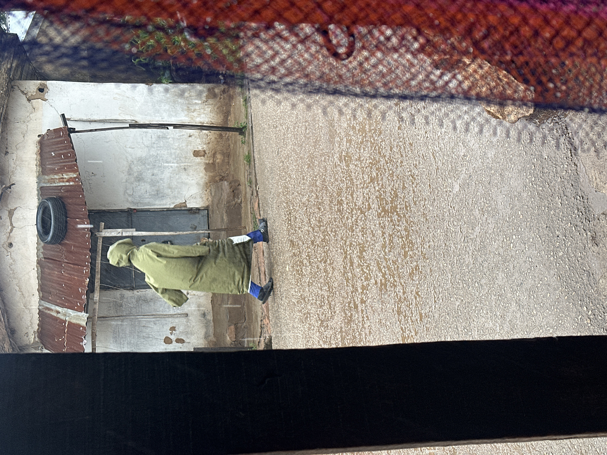 A person wearing a hooded, green, full-length coat and blue pants walks on a wet, asphalt road in front of a dilapidated building with a corrugated metal roof. The road is reflective and shows evidence of standing water, indicating it might be raining or has recently rained. The building's walls are white and textured, with some peeling plaster. A tire is resting on the roof of the building. The scene is framed on the left by a dark, vertical object and on the right by a reddish, textured material, possibly a curtain or net. The overall impression is one of a casual, everyday scene in what appears to be a less developed urban or rural environment.