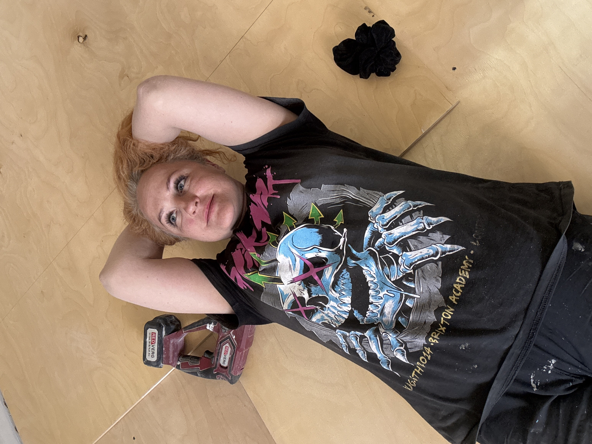 A woman with reddish-blonde hair is lying on a wooden surface, possibly a floor under construction or renovation. She is wearing a black T-shirt with a graphic print featuring a skull and the words "UGITH1014 BRIXTON ACADEMY". Her arms are behind her head, and she is looking at the camera with a slight smile.
Next to her is a red and black power tool, and further up is a black scrunchie.
There is no visual information in the image to indicate that it was taken in Chaikovsky, Russia.