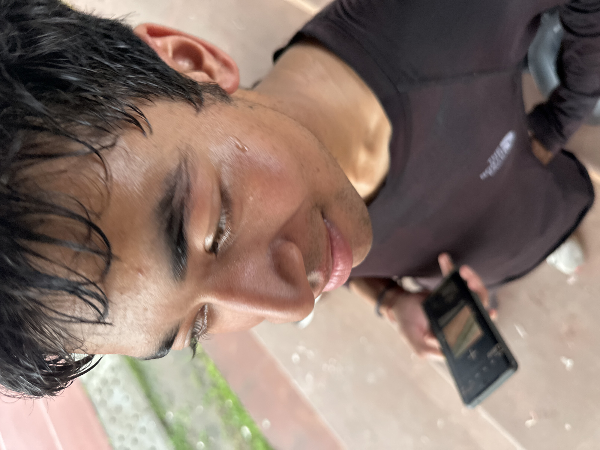 A young man with dark, damp hair looks down and to his left, holding a dark smartphone in his right hand. His skin appears slightly sweaty, with a visible bead of sweat on his cheek. He is wearing a dark t-shirt, and a bracelet is visible on his left wrist. The background shows a textured, reddish-brown surface, possibly bleachers or a walkway, with blurred green foliage in the distance. The lighting suggests an outdoor, possibly overcast, environment.