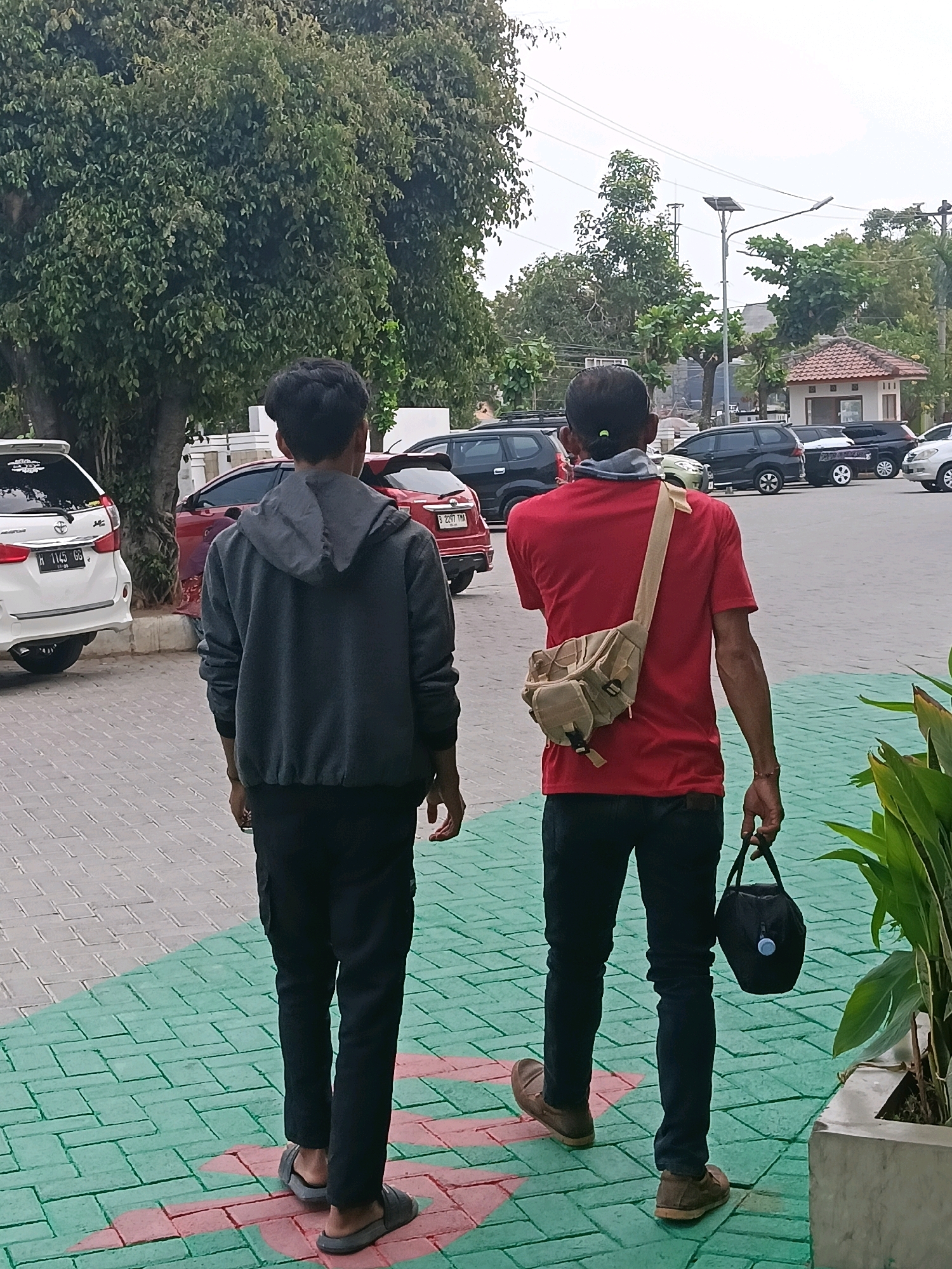 Two young men are walking away from the camera on a patterned brick sidewalk. The man on the left wears a dark gray hooded jacket and black pants. The man on the right wears a red t-shirt, dark pants, and has a beige shoulder bag. He is carrying a black bag in his right hand. In the background, several cars are parked in a lot, including a white SUV on the left and red and black SUVs to the right. There are trees and buildings visible in the distance. The scene appears to be an outdoor area in the city of Balun, Indonesia, possibly a parking lot or public space.