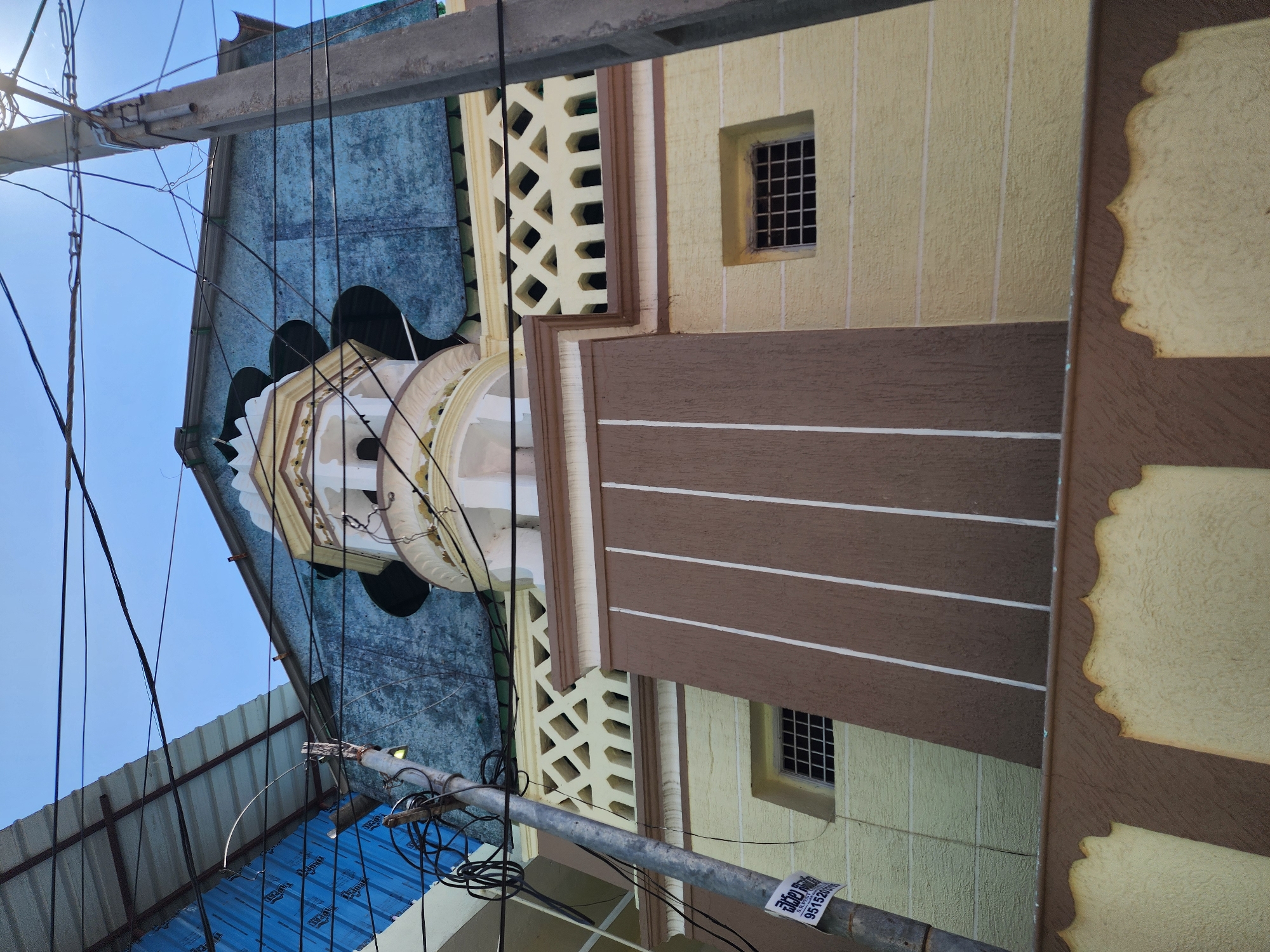 The image displays an upward view of a multi-story building facade in Pedda Kammavaripalli, India, against a clear blue sky. A concrete utility pole occupies the left side of the frame, laden with numerous black electrical and communication wires that crisscross the visual field. A yellow and white paper sign, bearing Telugu script and the number "95152007", is affixed to the pole.
The building's lower section features textured pale yellow walls. A central brown-painted area on the lower facade is vertically striped with three white lines. Two small, rectangular windows with black metal security grilles are observed within the pale yellow sections. Above this, a decorative pale yellow railing with diamond and rectangular openwork patterns is present.
A prominent, ornate tower-like structure, primarily white and pale yellow with arched openings and multiple tiers, extends upwards from the building's central section. The uppermost visible part of the building consists of a textured, weathered blue-green wall beneath a dark, possibly corrugated, roof edge. To the far left, a corrugated metal roof covered with blue material from an adjacent structure is partially visible. No individuals or specific actions are discernible within the frame.