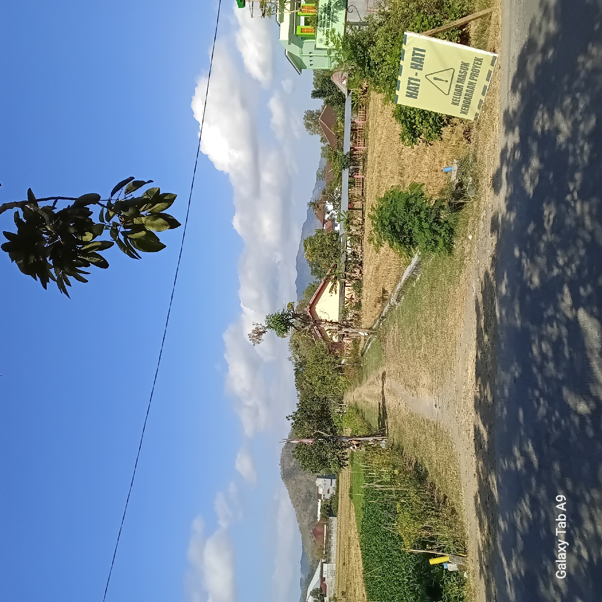 The image depicts a rural scene in Sambirejo, Indonesia, under a clear blue sky with scattered clouds. In the foreground, a paved road is visible, with dappled shadows cast by overhead foliage. To the right of the road, a bright yellow sign with black lettering and a warning symbol stands propped up. The text on the sign reads "HATI-HATI" (which translates to "Be Careful") and "KELUAR MASUK KENDARAAN PROYEK" (which translates to "Project Vehicle Entry and Exit").
Beyond the road, a dirt path leads into the distance, flanked by agricultural fields. On the left, a field of green crops is visible, while the field on the right appears to be a harvested dry crop, possibly rice. Further into the background, various buildings and structures are scattered amongst trees. These include a yellow building with a red roof, a modern-looking green building with signage that indicates it might be a government office or facility, and other residential or agricultural buildings. In the far distance, rolling hills form the horizon. A tree with dark green leaves hangs into the frame from the top right corner. The overall impression is one of a sunny day in a developing rural area.