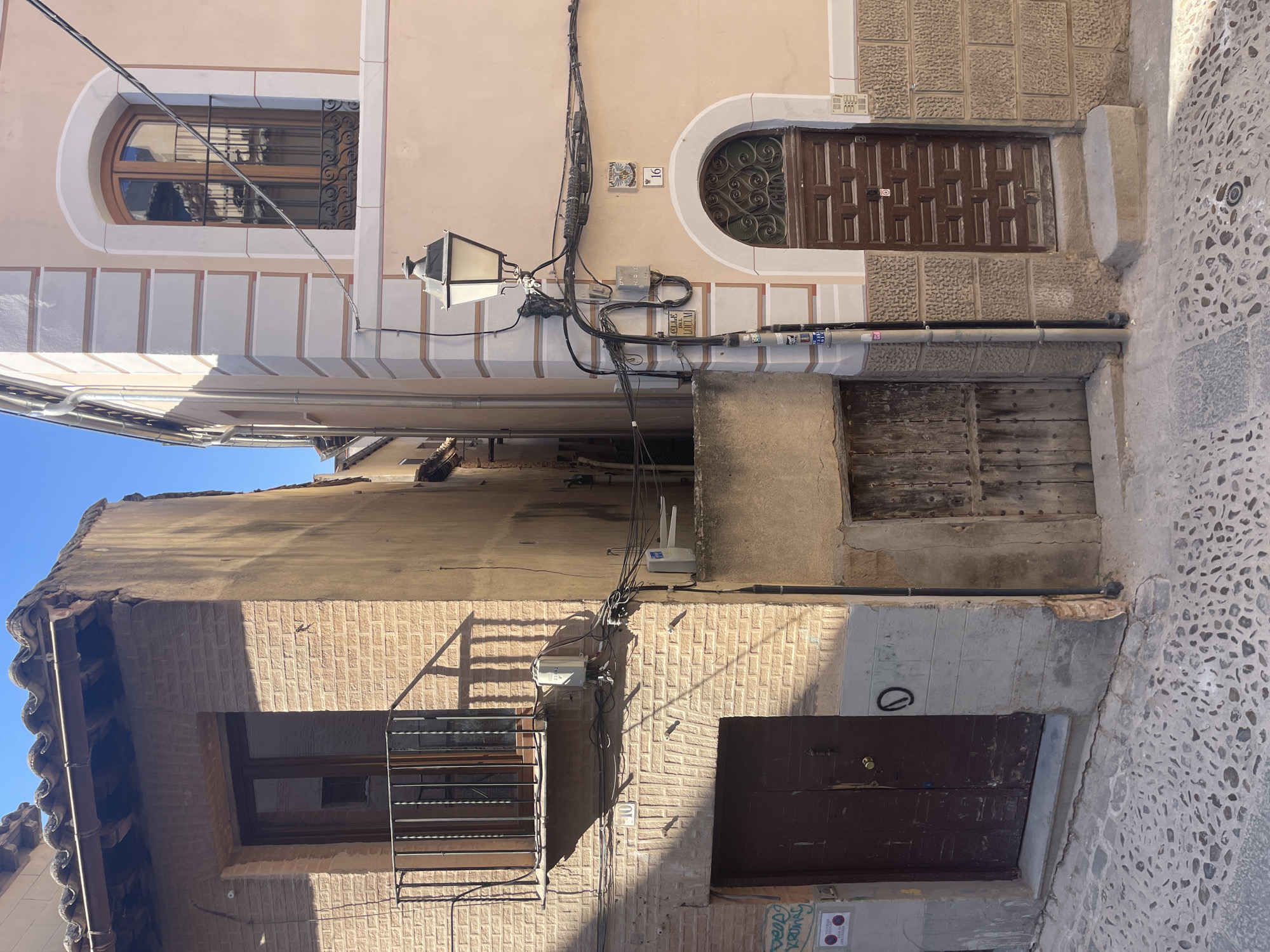 The image depicts a narrow cobblestone street in the city of Toledo, Spain, with two buildings meeting at a corner. The building on the left is constructed of light-colored brick and features a dark wooden door at street level and a balcony with a black metal railing above. The building on the right is painted a pale peach color and has decorative light beige trim around a large arched window with a wooden frame. A dark wooden door with decorative metalwork above it is visible on this building. A traditional street lamp with a black metal post and decorative scrolled arms is positioned between the two buildings. Various electrical conduits and junction boxes are attached to the facades of both structures. The sky above is clear and blue.