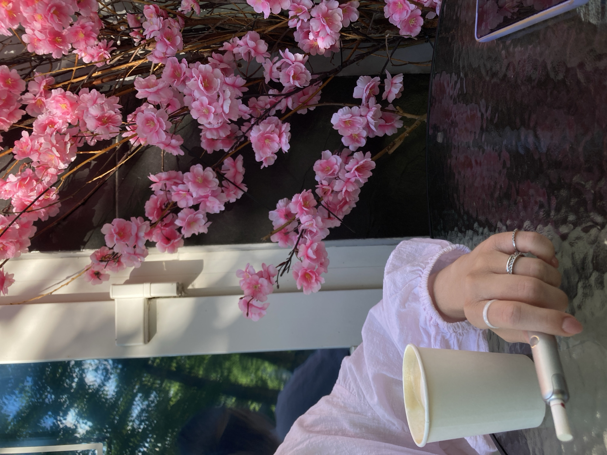 This media file features a close-up shot of a person's hand holding a white cup and a white cylindrical object, possibly an electronic cigarette or vape pen. The hand is adorned with multiple rings, including a silver band with a textured pattern and a thinner, plain silver ring. The person is wearing a white blouse with ruffled sleeves.
In the background, artificial pink cherry blossoms are arranged on branches, creating a decorative element. The setting appears to be outdoors, with a glass table visible and reflections of trees and sky on its surface, suggesting a daytime scene. There is also a white window frame with a hinge. The overall mood is relaxed and casual. No visible text is present in the image. The location context is Chaikovsky, Russia.