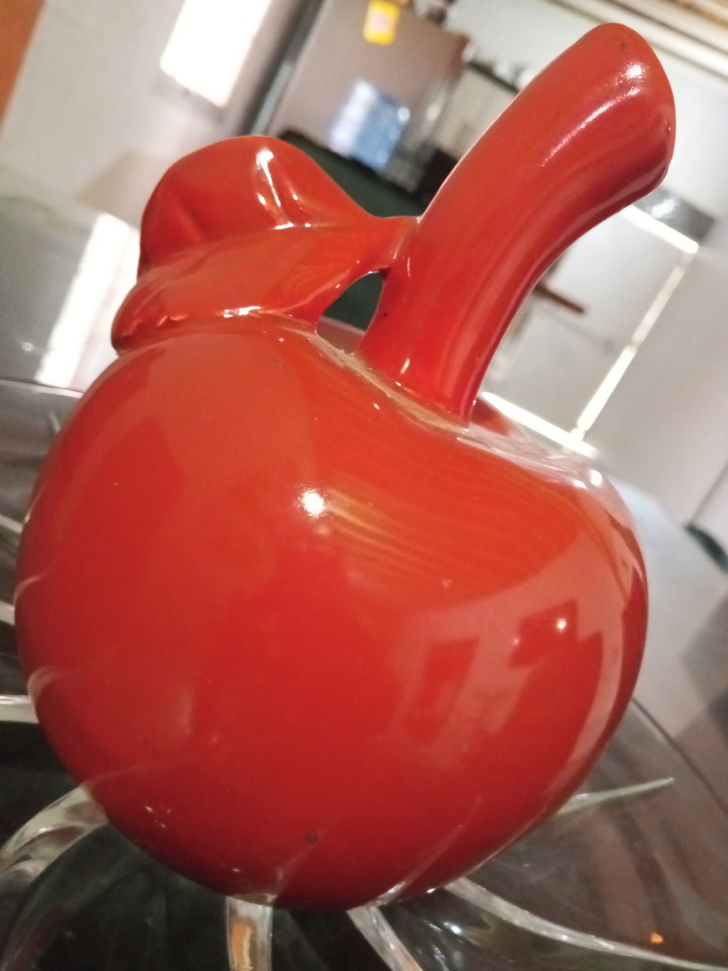 A close-up shot of a glossy, red ceramic apple figurine rests on a reflective glass surface, possibly a tabletop. The apple features a stem and a stylized bow-like detail near the top. The background shows a blurry interior setting, possibly a kitchen or dining area, with glimpses of appliances and other objects. There is no specific information about the location in the city of Altagracia, Venezuela, or details about people, actions, or interactions.