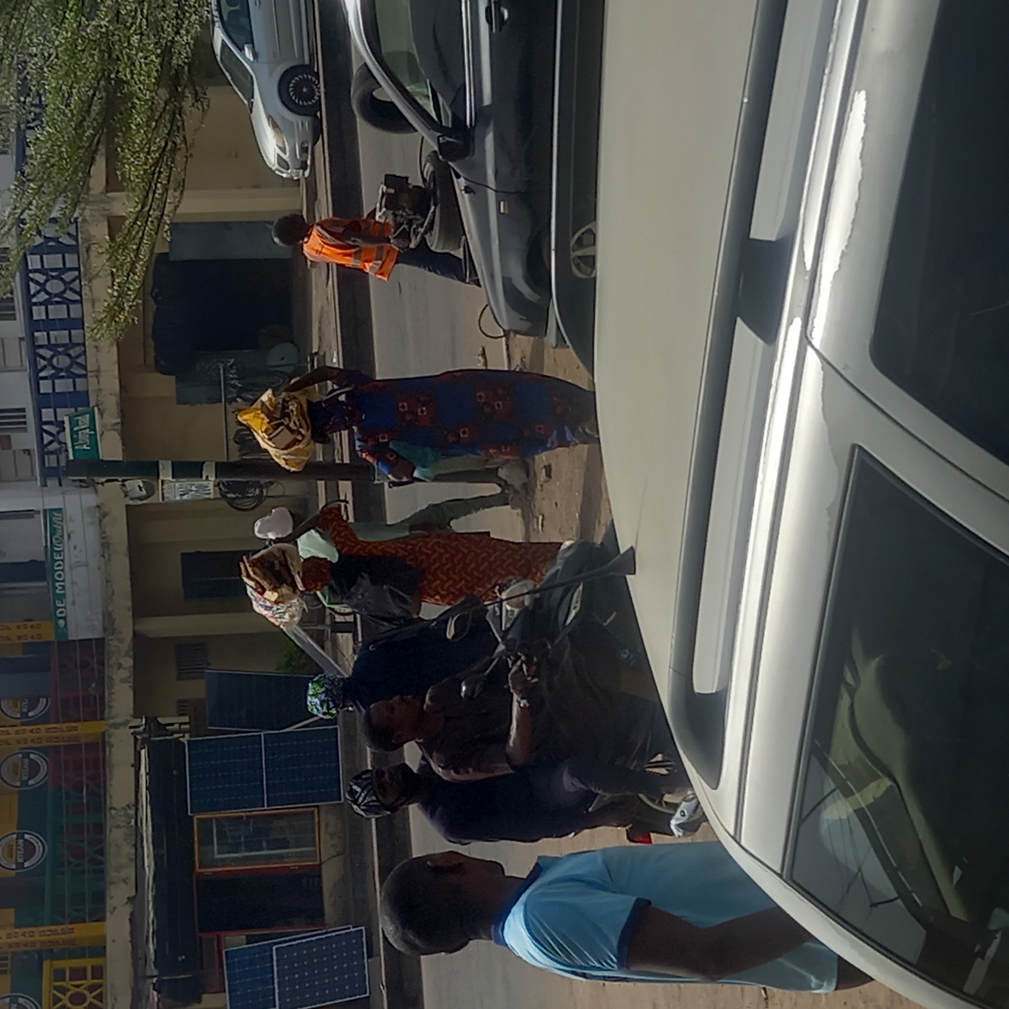 This daytime street scene captures a lively urban or semi-urban environment. In the immediate foreground, the side and roof of a white sedan are visible on the right. To its left, a man in a light blue polo shirt stands, seen from behind and side, looking towards the right.
Behind him, two men are on a motorcycle or scooter; the driver wears a helmet. Further left, a man in an orange safety vest and dark trousers is standing near a dark-colored vehicle, possibly a car or SUV. Several women are walking along the street; one woman in a blue patterned dress carries a yellow bag on her head, while another in a red patterned dress also carries items.
Buildings line the street in the background. On the left, several solar panels are propped against a structure. Above these, stacked commercial crates are visible, some bearing text like "KOLA" and "COCOA". Another building features a green sign that reads "DE MODE OUTFIT". A street pole in the mid-ground displays a smaller green sign with "Akinyemi Rd" written on it. Further in the background, another white car is parked. Trees with green foliage are visible on the far left. The scene depicts a bustling street with daily activities, pedestrians, vehicles, and commercial elements under bright daylight.