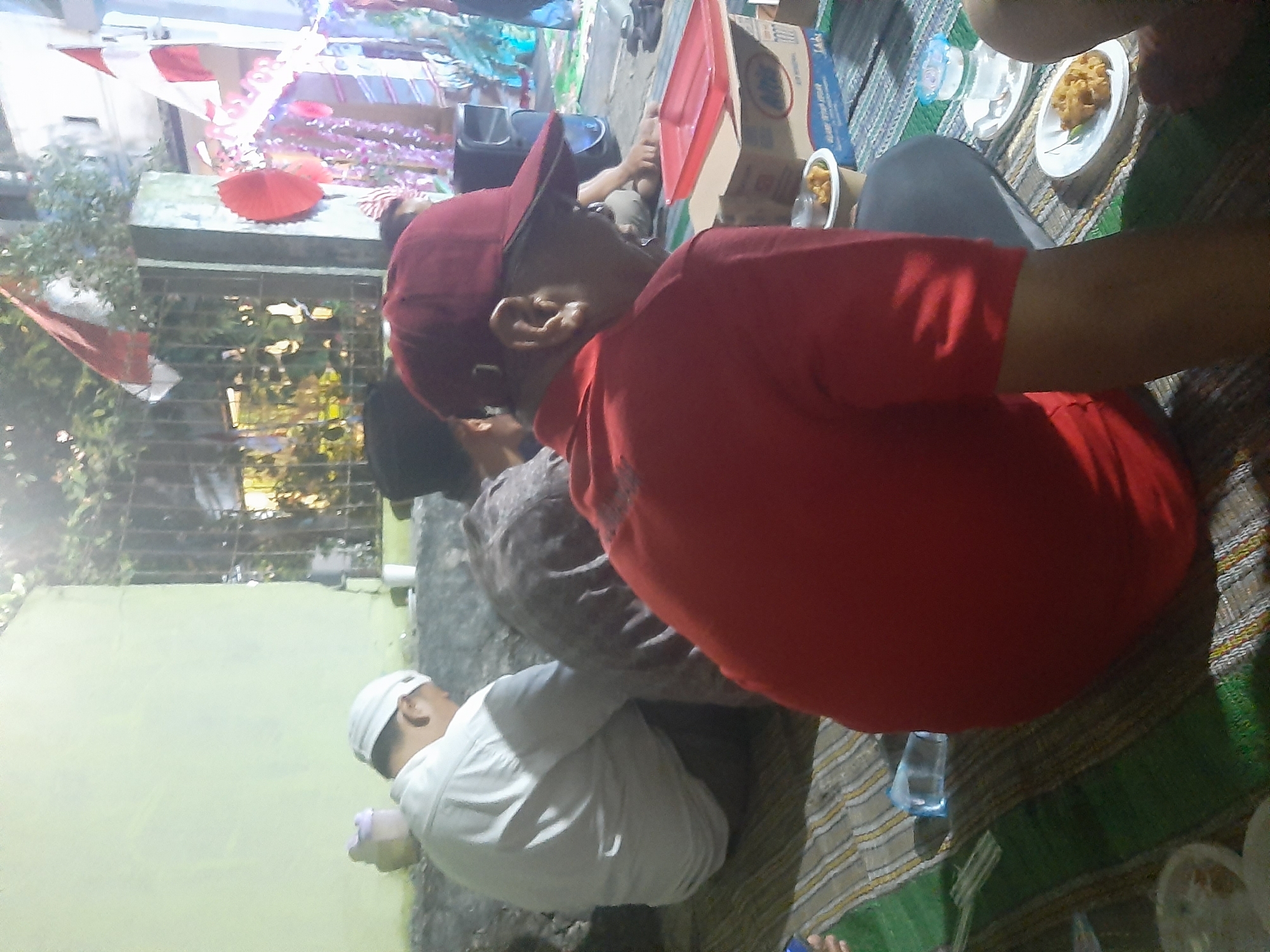 In the city of Sumampir, Indonesia, an outdoor gathering is underway. The scene is viewed from a low angle, focusing on three individuals seated on the ground. In the foreground, a person wearing a red shirt and a red baseball cap is partially visible, their right ear and side of their face in profile. Behind them, a second individual in a white shirt and white cap is seen from the back, their head bowed. To the left of the person in white, another person is seated, their form obscured by a dark, patterned cloth. The ground is covered with striped mats in shades of green, white, and brown. In the background, a building with a light green wall is visible, adjacent to a metal fence. Beyond the fence, trees and foliage can be seen, along with decorative elements including red circular ornaments and streamers. A black speaker is also present in the background, suggesting music or a public address system. On the ground, several items are scattered, including a red plastic tray, a cardboard box with blue markings, a small white cup with a white lid, and plates with food. A clear plastic bottle with a blue cap is also visible. The lighting suggests it is daytime, with some light filtering in from the upper right of the frame.
