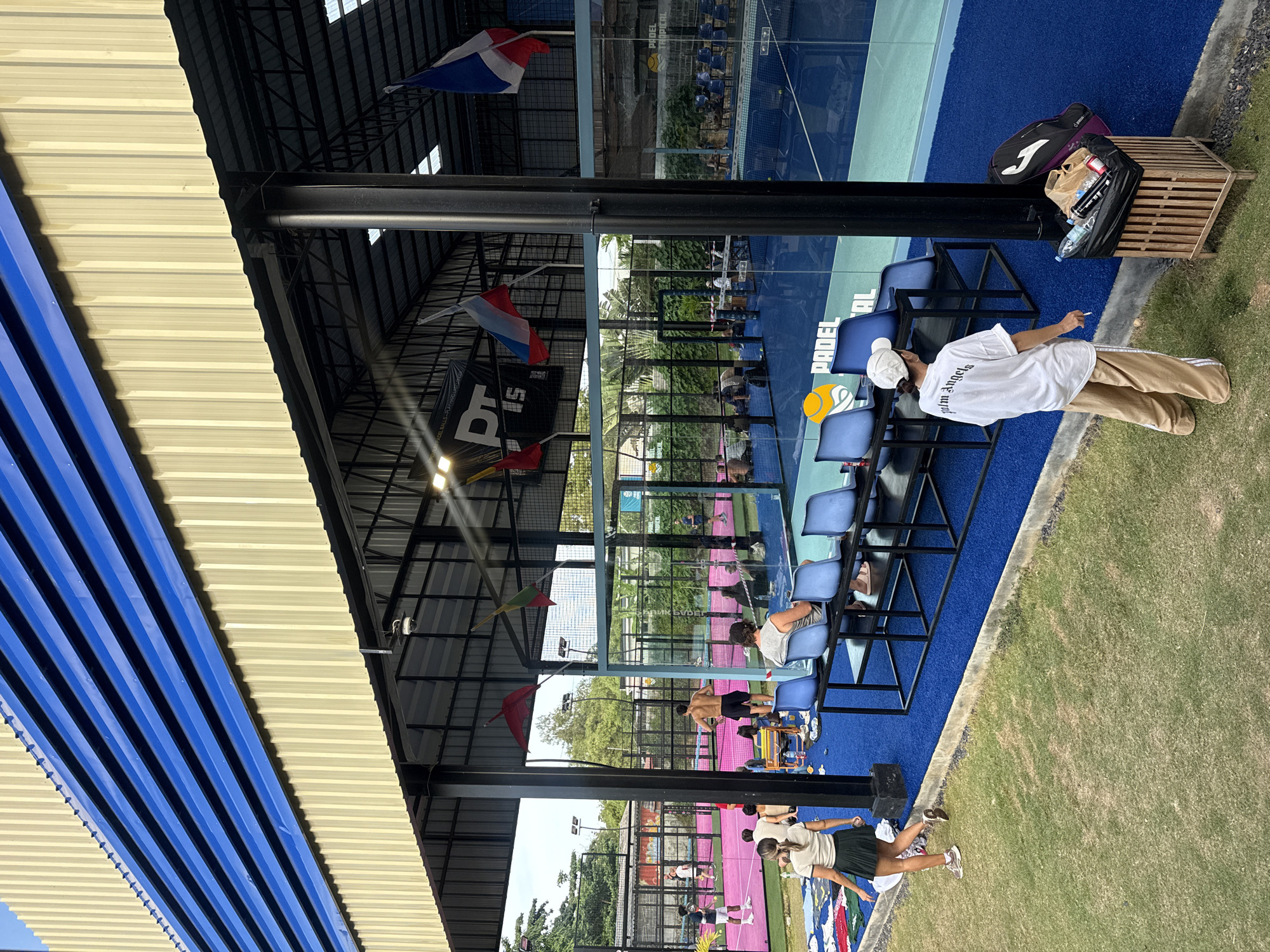 The image shows an outdoor padel tennis facility in the Ko Samui District of Thailand. In the foreground, a woman in a white t-shirt and khaki pants stands facing away from the camera, holding a cigarette. A trash can filled with bottles and bags sits next to her. Behind her, on the grass, are various items of clothing.
The facility features several padel courts with blue and pink artificial turf. Spectator seating, consisting of blue chairs on black metal stands, is visible under a covered area with a corrugated metal roof. Several flags are hung from the structure, including what appear to be the flags of Belgium, Russia, and France. People are seen on and around the courts, some playing padel and others observing. The overall environment appears to be a recreational sports complex with lush greenery surrounding the courts.