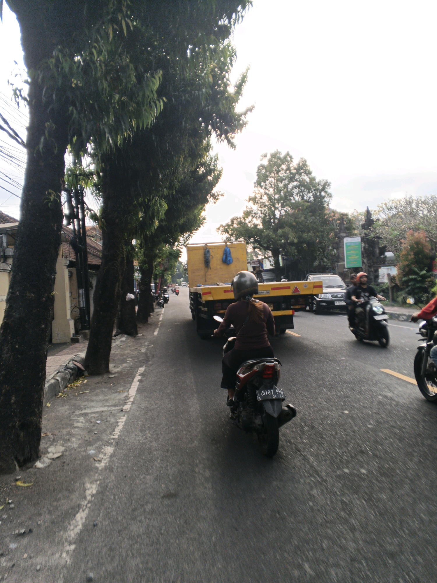 The image captures a street scene in Blahbatuh, Indonesia, on a cloudy day. In the foreground, a person rides a scooter with a license plate visible that reads "L 5197 PV" and "01-22". They are wearing a brown shirt and a silver helmet. Behind them, a yellow flatbed truck with a blue tarp is stationary. To the right, another scooter rider, wearing an orange helmet, is in motion. Further down the road, more scooters and a black car are visible. The street is lined with large trees on the left, some with moss growing on their bark. Sidewalks are present with some debris and fallen leaves. Buildings can be seen along the left side of the road. The overall atmosphere suggests a typical day of traffic and activity in a suburban or urban area.