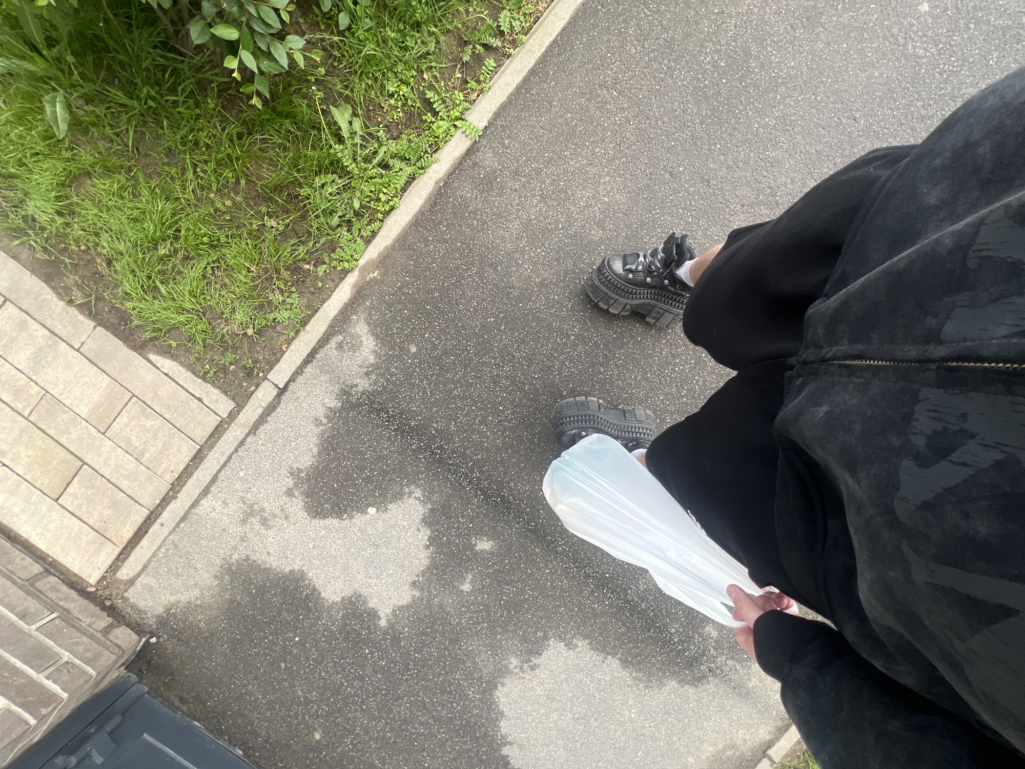 The image shows a person walking on a wet asphalt sidewalk next to a brick wall and some grass. The person is wearing black shorts and a black zip-up hoodie. They are carrying a translucent white plastic bag in their right hand. The person is wearing chunky black platform boots with laces. The location is a street in Saint Petersburg, Russia, indicated by the architectural style of the brickwork and the presence of a typical Russian sidewalk. The weather appears to be overcast or recently rained.