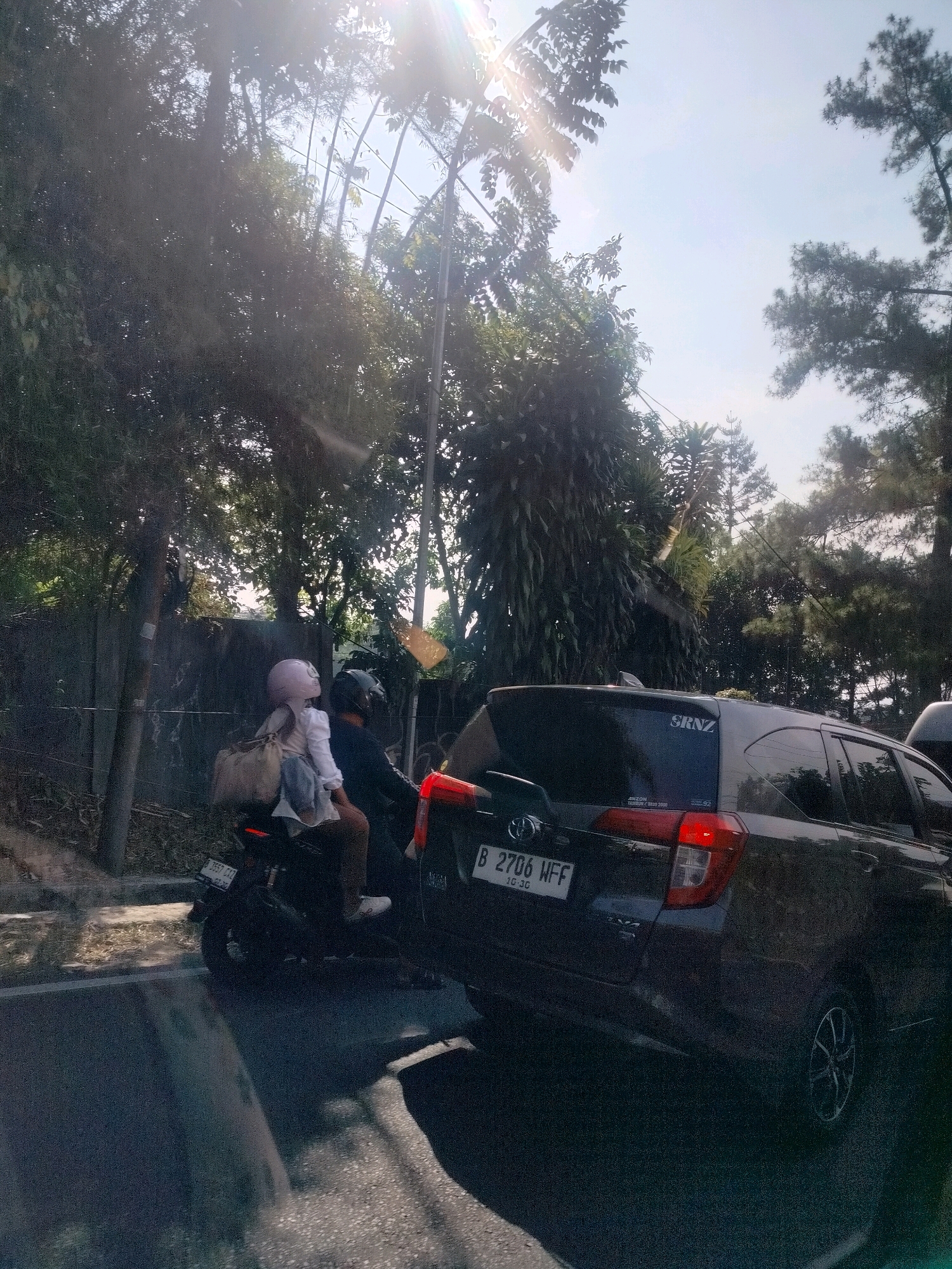 The image depicts a street scene in Cisarua, Indonesia, on a sunny day. In the foreground, a dark-colored car is visible, its rear end facing the camera. The car has a license plate that reads "B 2706 WFF" and a sticker on the rear window says "SRNZ". To the left of the car, a motorcycle is seen from the side. Two people are riding the motorcycle; a person in front is wearing a white shirt and a pink helmet, and a person behind is wearing a dark helmet and dark clothing. The motorcycle has a license plate that reads "B 3557 CXJ". The road is paved, and there is a concrete barrier or wall to the left of the road, lined with trees and vegetation. The background is dominated by lush green trees and a bright, sunlit sky. The overall impression is a snapshot of daily life in a tropical urban environment, with traffic and natural elements coexisting.