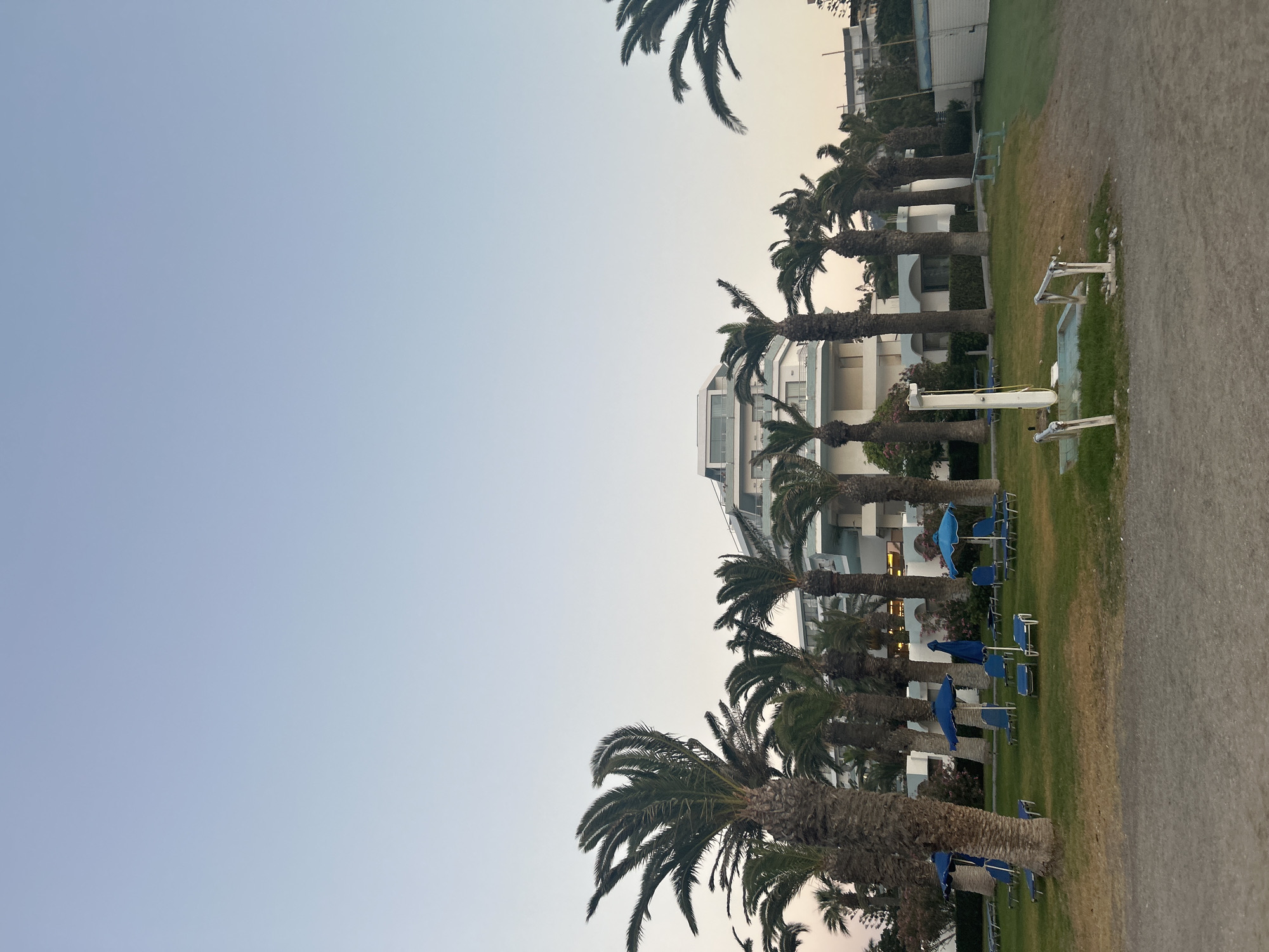 The image depicts a resort scene in Rhodes, Greece. A modern, multi-story hotel with white balconies and a distinctive angular roof is visible in the background. Palm trees are scattered throughout the property, with some in the foreground casting shadows on a well-maintained lawn. Several blue lounge chairs and umbrellas are set up on the grass, suggesting a recreational area. A gravel path runs in front of the lawn. The sky is a clear, pale blue, indicating either early morning or late afternoon. No people are visible in the image.