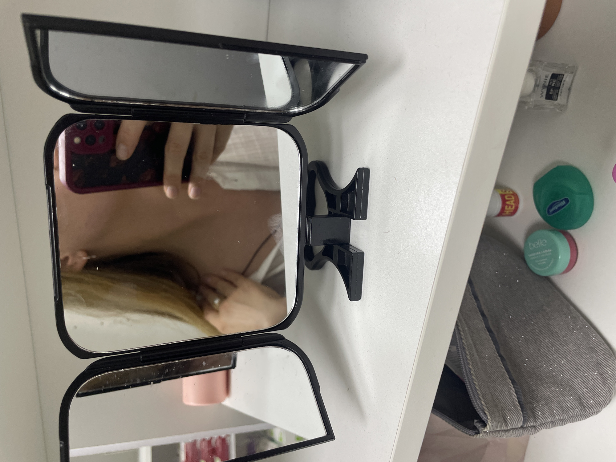 An indoor, eye-level shot shows a person holding a mobile phone and taking a selfie in a tri-fold mirror. The mirror is black and sits on a white surface, possibly a shelf. The person's face is visible in the center and right panels of the mirror, with their right hand holding a red-cased phone in the center panel. The left panel of the mirror reflects a pink cylinder and a white shelf with multiple compartments. Below the white surface, a grey, zippered makeup bag with a glittery texture is visible, along with two small containers of beauty products: one green and one pink. A small clear bottle with a black label is also present. The overall scene suggests a personal grooming area.