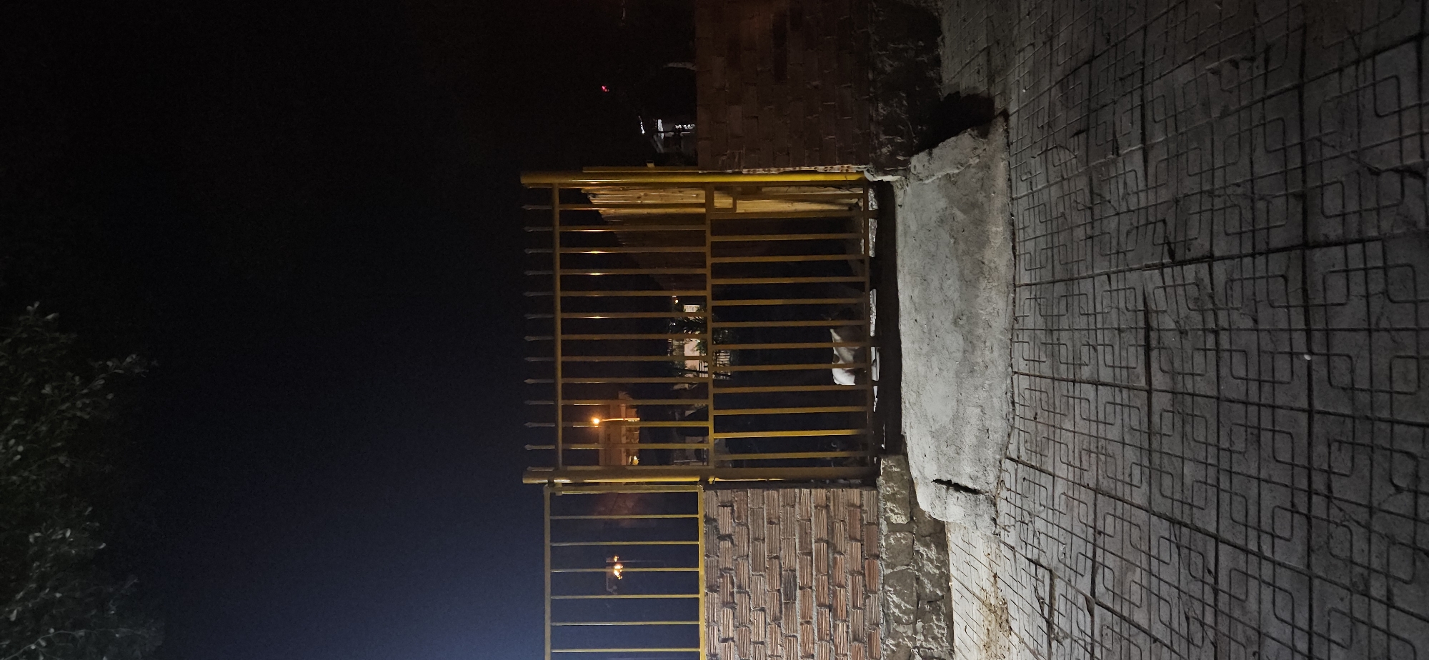The image captures an outdoor scene at night, likely in Bac Nha Trang Ward, Vietnam, with a strong focus on a yellow metal gate, a brick wall, and a patterned concrete pathway.
The central subject is a tall, yellow metal gate, constructed from numerous horizontal bars, appearing to be an entrance or part of a boundary wall. To the right of the gate, a section of a reddish-brown brick wall is visible, exhibiting a stacked pattern. Further to the right and extending into the foreground is a concrete path or ground, characterized by an embossed, grid-like pattern made of interconnected square or loop shapes.
Through the bars of the yellow gate, hints of artificial lighting can be seen in the distance, including a distinct bright orange-yellow light source. Behind the gate, a small, white-colored animal, possibly a cat or a small dog, is partially visible. On the far left edge of the frame, dark foliage or tree branches are dimly discernible against the dark background.
The time of day is clearly night, as the surroundings are enveloped in deep shadow, and artificial lights are the only significant illumination. There is no discernible human activity or specific event taking place. The weather conditions cannot be determined beyond the clear night sky. No text is visible in the image.