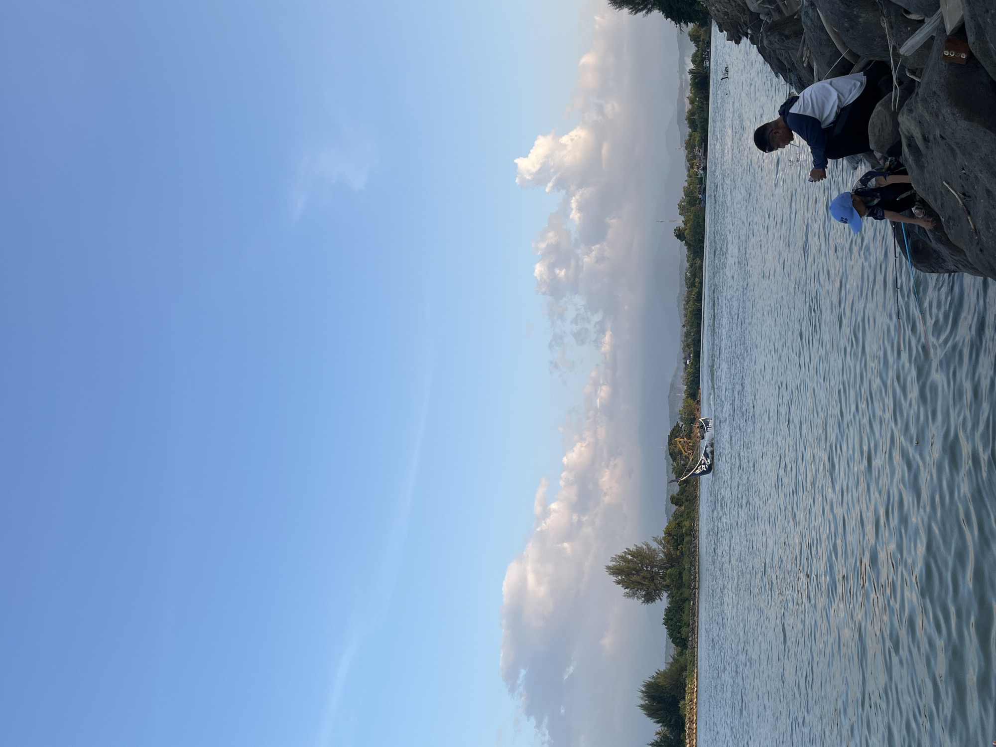 An outdoor scene depicts a body of water with a rocky shore. Two individuals, an adult male and a child, are seated on the rocks near the water's edge. The adult is wearing a white and blue jacket and dark pants, and appears to be smoking. The child is wearing a blue hat and a dark patterned shirt. They are both looking towards the water. In the distance, across the water, is a treeline with some vegetation and hills beyond. A boat with a dark sail is visible on the water in the mid-ground. The sky is clear with some scattered white clouds, and the lighting suggests it is late afternoon or early evening. The scene is located in Meunasah Jurong, Indonesia.