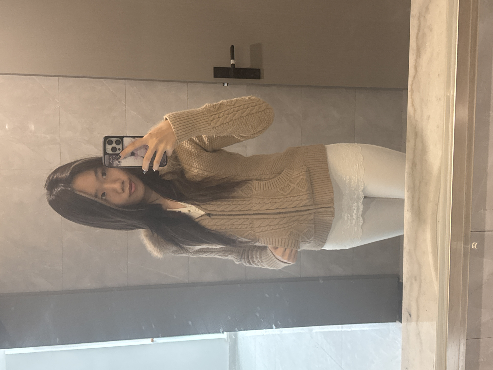 The image displays a young East Asian female, depicted in a mirror selfie indoors. She has long, dark hair with lighter highlights. The individual is wearing a light brown cable-knit hooded cardigan with a visible fur-like lining around the hood, a white collared top partially visible underneath, and white leggings with a white lace trim at the upper thigh. She holds a dark-colored smartphone, featuring a multi-lens camera system and a patterned case, in her right hand, oriented towards the mirror. Her left hand is placed inside a pocket of her cardigan. The environment, reflected in the mirror, consists of light grey tiled walls, a light-colored countertop in the foreground, and a plain light brown door with a black handle on the right. A section of a light-colored wall with a dark vertical strip is visible on the far left. The specific location within Nan'an District, China, cannot be determined from the provided image.