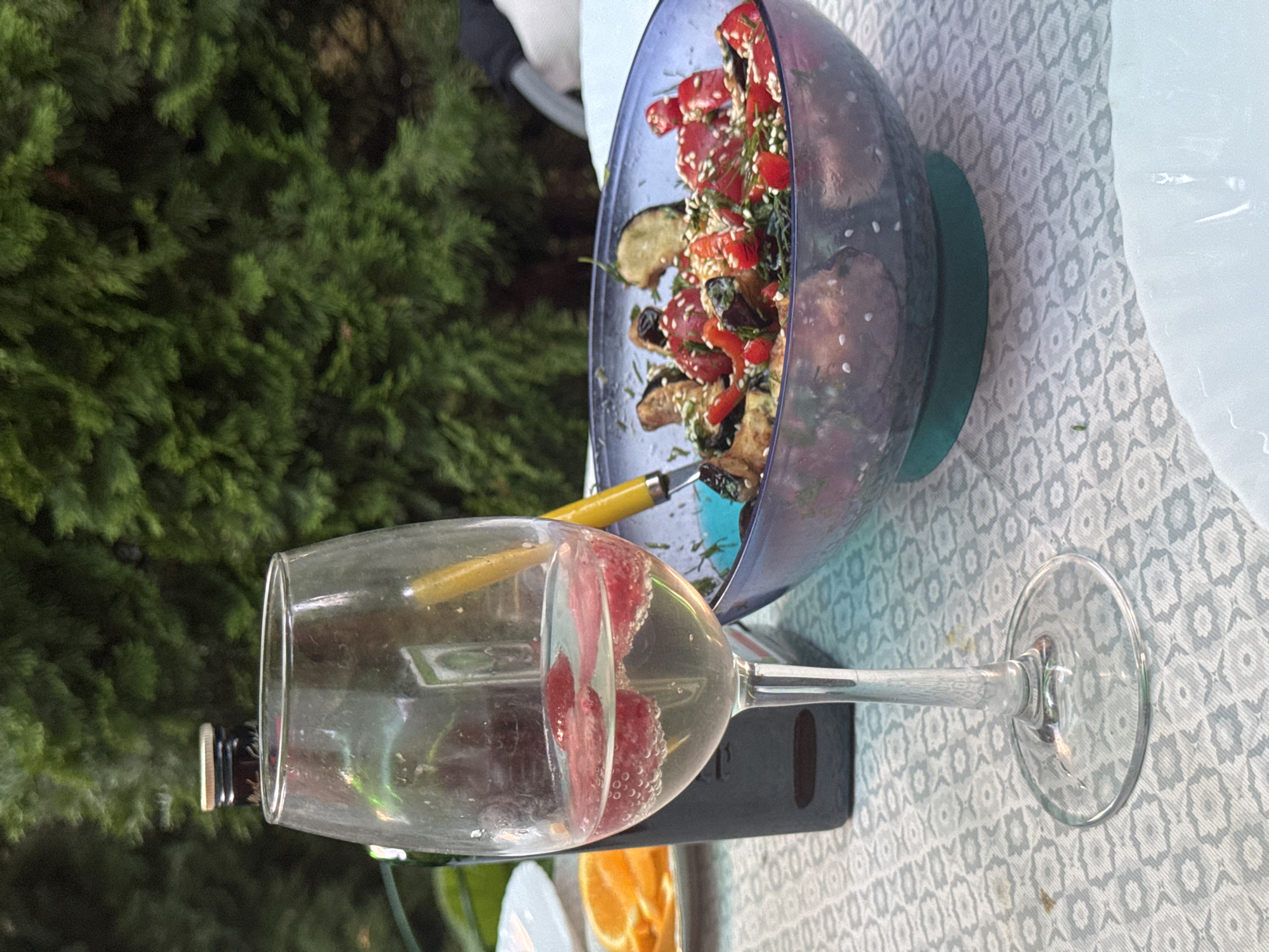 A glass of white wine with strawberry slices sits on a table covered with a patterned tablecloth. To the right of the glass is a blue bowl filled with a salad of roasted vegetables, including eggplant, tomatoes, and peppers, garnished with sesame seeds and dill. Behind the wine glass, a dark bottle, possibly olive oil or wine, is visible. In the background, lush green foliage suggests an outdoor setting, possibly a garden or patio. A white plate with orange slices is partially visible to the left of the wine glass. The scene is likely located in Pribrezhnyi, Russia, given the context.