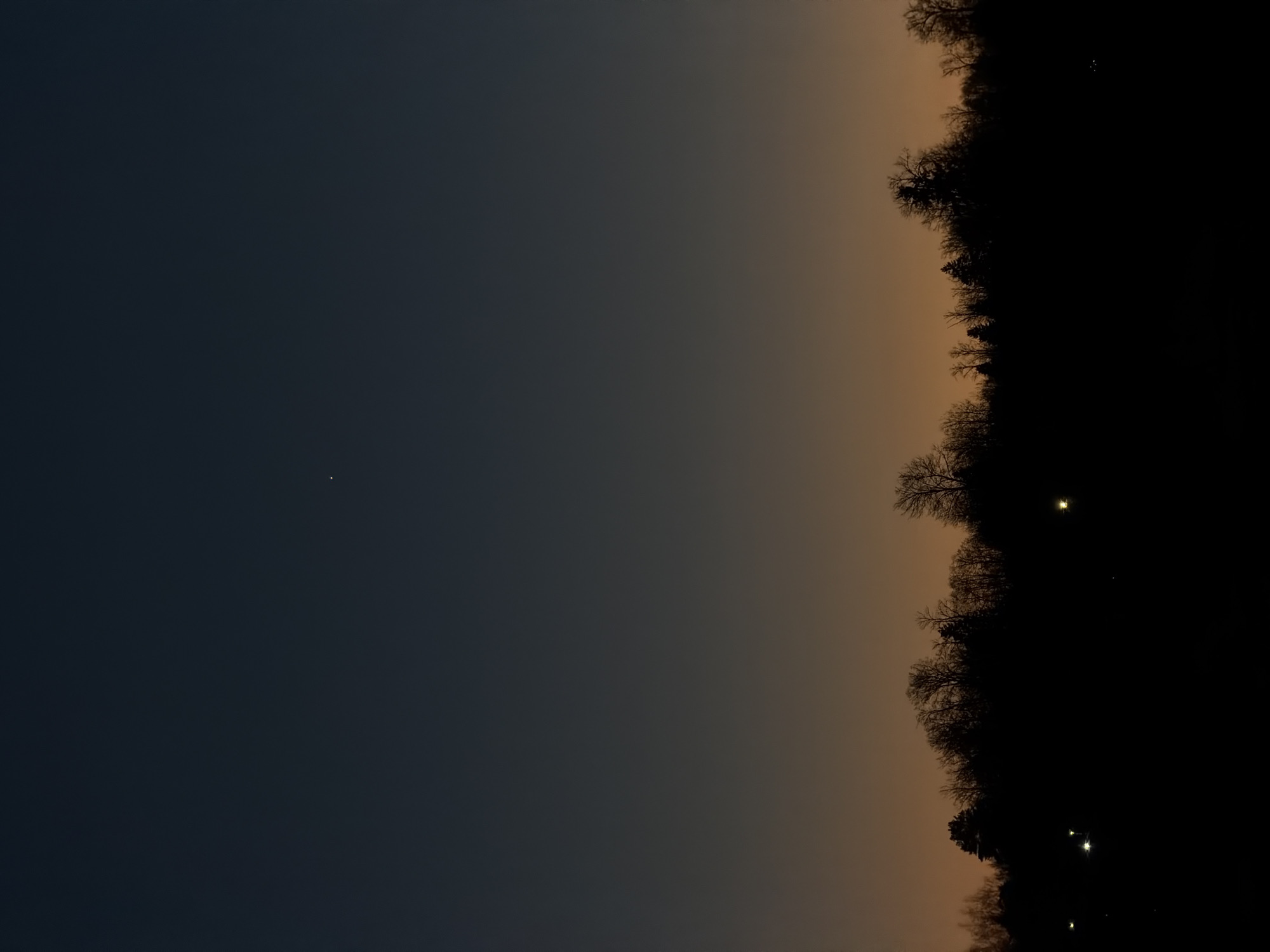 The image shows a night scene in what could be a rural area near Izhevsk, Russia. The sky is a gradient, dark blue on the left fading to a light orange-brown towards the horizon. A single bright star is visible in the dark blue portion of the sky. The right side of the image is dominated by a dark silhouette of trees, likely a forest or a dense group of trees. There are a few small points of light interspersed within the trees, possibly distant streetlights or house lights. The photo appears to be taken on the outskirts of Izhevsk, where urban lighting meets natural landscape.