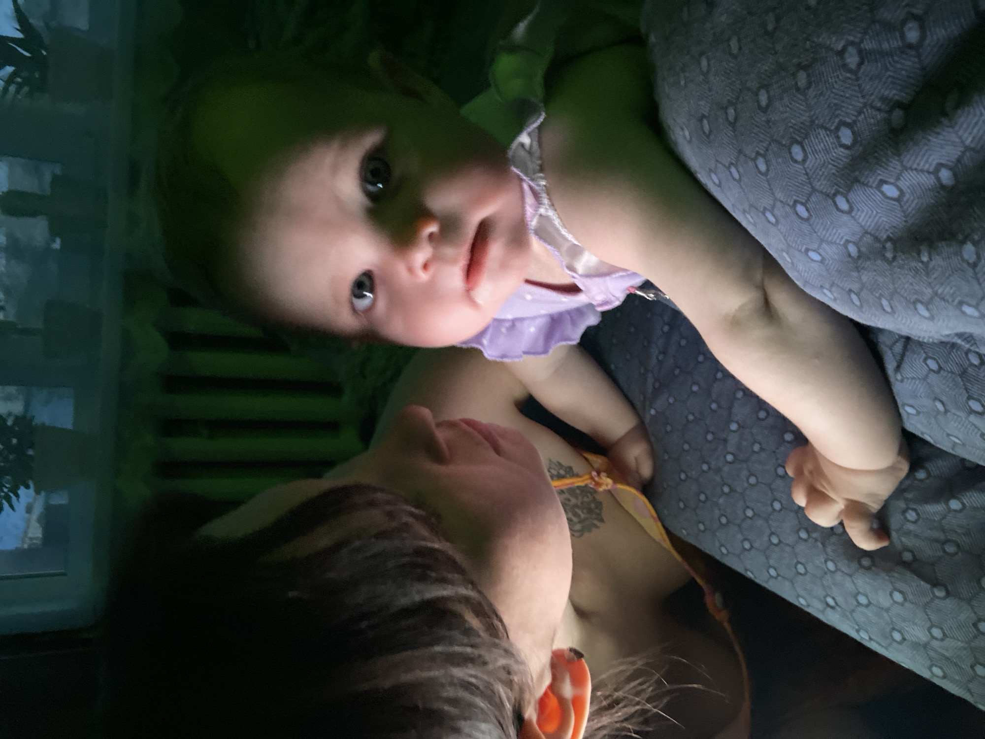 A baby wearing a purple and white polka-dot dress lies on a patterned gray blanket while held by a woman. The woman has short brown hair, and a tattoo on her upper chest/shoulder area. The lighting is dim with greenish hues. A window is visible in the lower left corner of the image, showing a dark exterior with some vague foliage. There is no indication that the location is the city of Фоки, Russia.