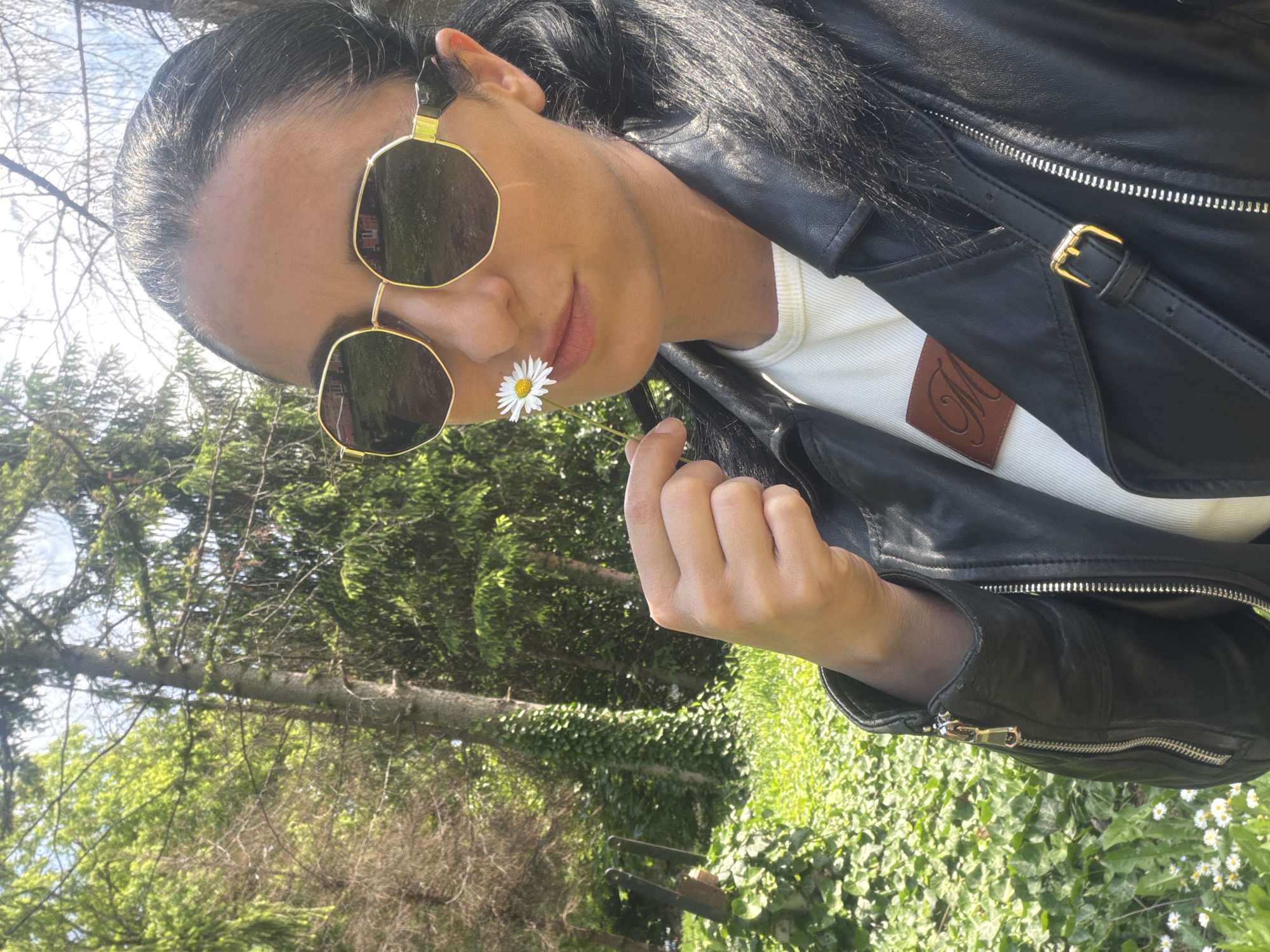 A woman is posing outdoors in a natural setting. She is wearing a black leather jacket over a white top, and sunglasses. She is holding a daisy near her nose. The background consists of green foliage, trees, and glimpses of the sky. The photo appears to be taken during the day with natural light. The image doesn't provide any information about the location in Pleven, Bulgaria.