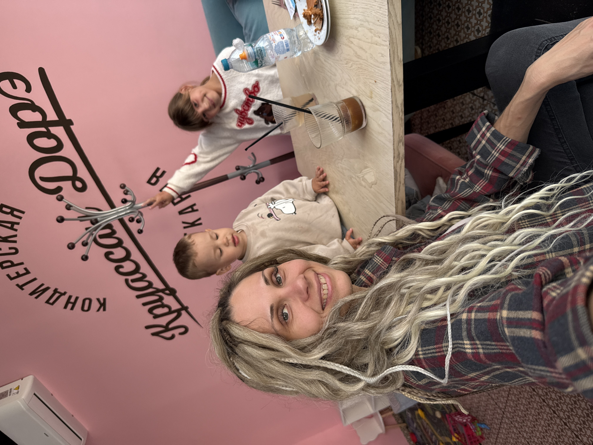 A selfie-style, canted-angle photograph shows a woman, a young girl, and a toddler seated at a table inside a "Kruassanitsa" café in Rusanovo, Russia. The woman is centered in the foreground, with long, blonde wavy hair that appears to have white braids. She is wearing a red and black plaid shirt and smiles at the camera. Behind her, a toddler in a light-colored sweater is sleeping. To the toddler's left, a young girl in a white sweater with a red heart design leans toward a decorative coat rack attached to a pink wall, which has black lettering that appears to be the café's name. The table is a light wood and features two glasses, straws, a plate with food, and a bottle of water. A person's arm in a plaid sleeve and dark pants is visible on the right side of the image. A decorative tile pattern is visible behind the woman on the floor. An air conditioning unit is visible on the wall below the decorative lettering.