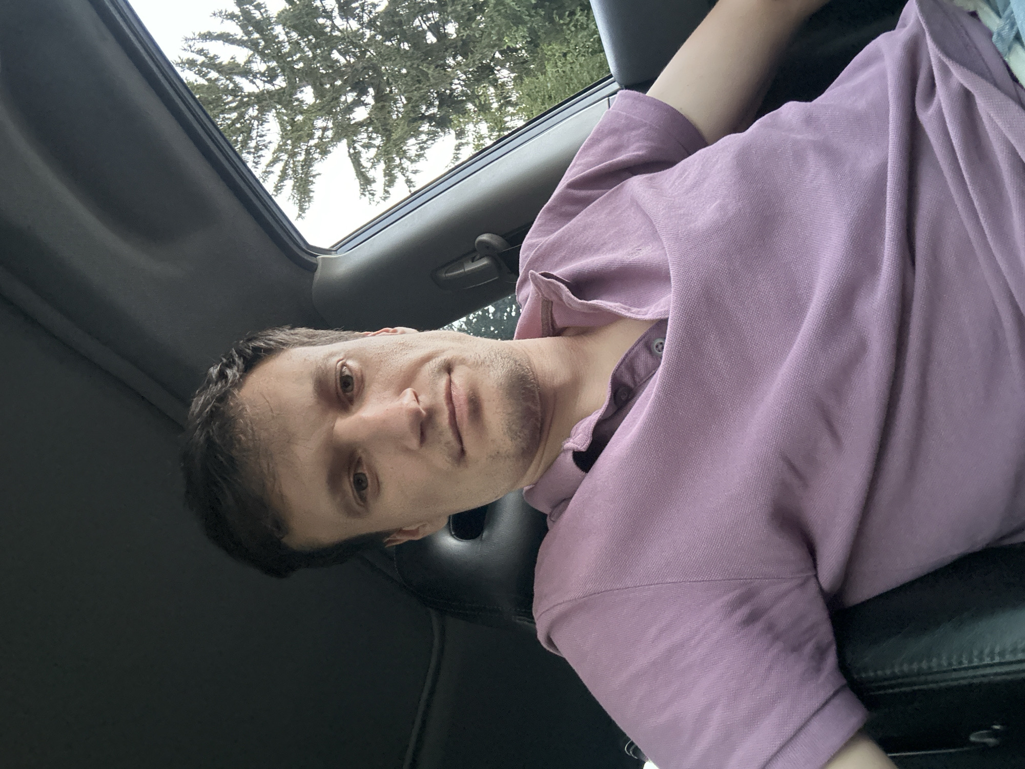The image shows a man in a purple polo shirt sitting in a car. He is looking at the camera. The car interior is visible, including a black leather seat and the dashboard. Outside the car window, a tall evergreen tree is visible against a cloudy sky. The man appears to be in his late 20s or early 30s with brown hair and a light beard. The image was taken in Shatava, Ukraine.
