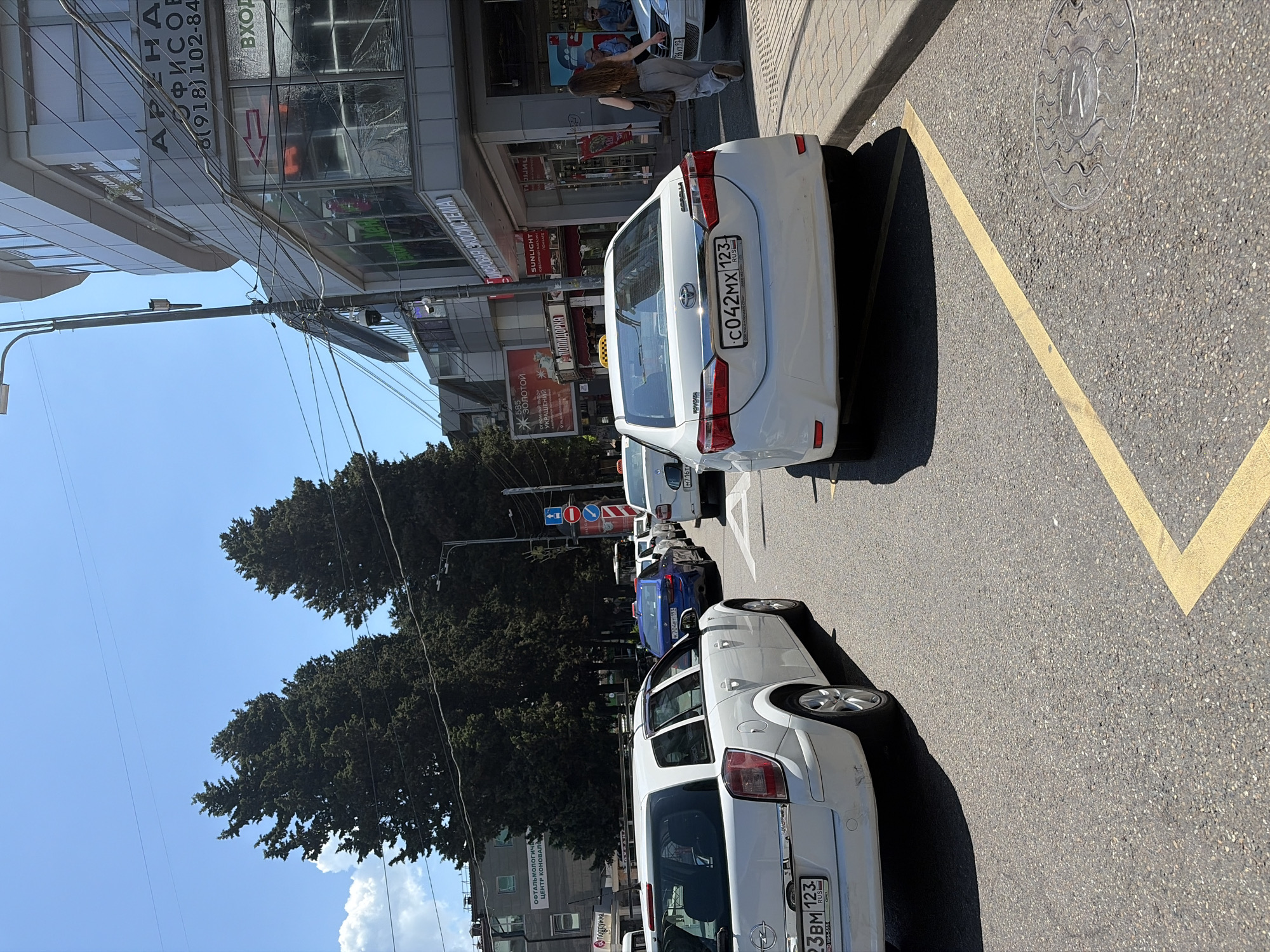 This is a street-level, medium shot photograph taken on a sunny day in Sochi, Russia. The scene depicts a busy street with several cars, including a white Opel Astra wagon in the foreground and a white Toyota Corolla taxi in the mid-ground. A woman with long brown hair is walking away from the Toyota Corolla towards a building on the right. The building appears to be a commercial establishment with signage that includes "Sunlight" and "Ювелирный магазин" (Jewelry store). In the background, there are tall green trees and a blue sky with some clouds. The street has yellow lane markings. Several traffic signs are visible, including a red arrow pointing left and traffic regulation signs. The overall impression is of an urban environment with ongoing traffic and pedestrian activity.