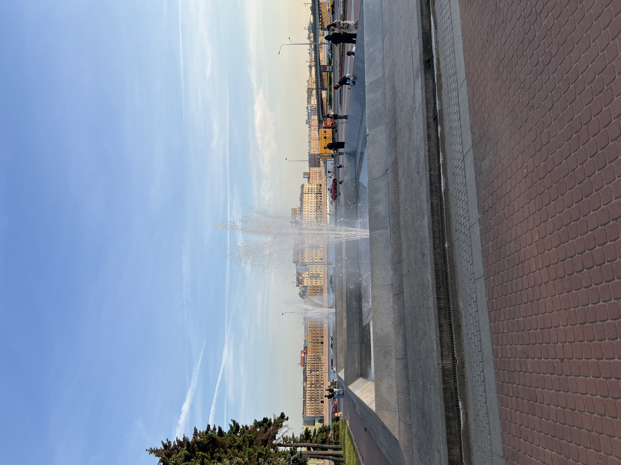 The image depicts a view of the Neva River embankment in Saint Petersburg, Russia, during daylight. In the foreground, a large concrete fountain with active water jets is visible. The fountain basin is constructed from multi-tiered concrete structures. Beyond the fountain, a paved walkway and green lawn lead towards the river.
Across the Neva River, a row of historic buildings with light-colored facades and yellow accents lines the opposite bank, suggesting a cityscape. A bridge spans the river in the middle ground. Several people are present on the embankment, some near the fountain and others walking or standing along the riverside. A red car and other vehicles are also visible on the road parallel to the river. The sky is blue with some light clouds and contrails. The overall scene conveys a public space, likely a park or promenade, in an urban setting.